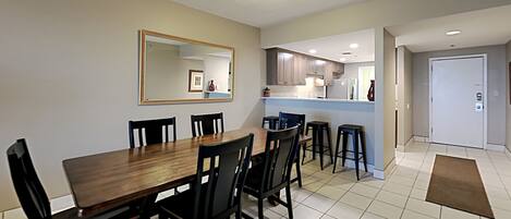 A dining area with a wooden table and chairs, a kitchen in the background, and a door leading to another room.