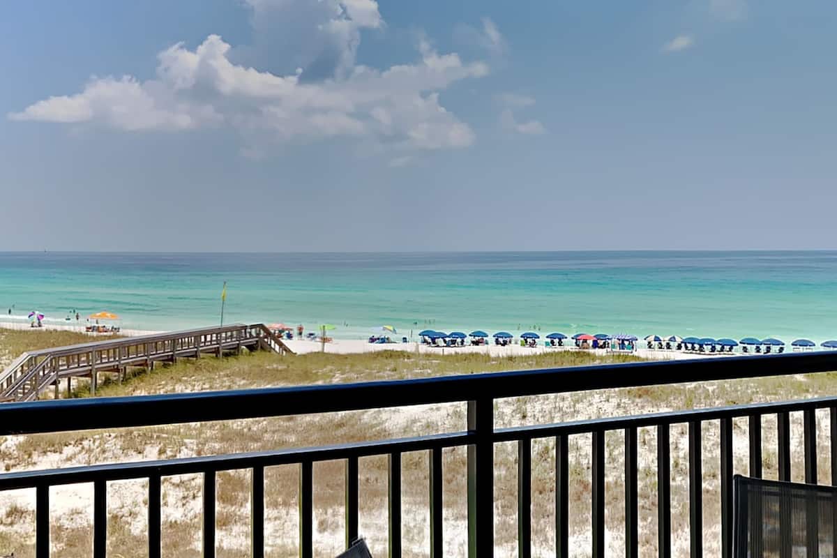 Une vue sur la plage avec une promenade en bois menant au rivage, bordée de parasols et de personnes se prélassant sur la plage.