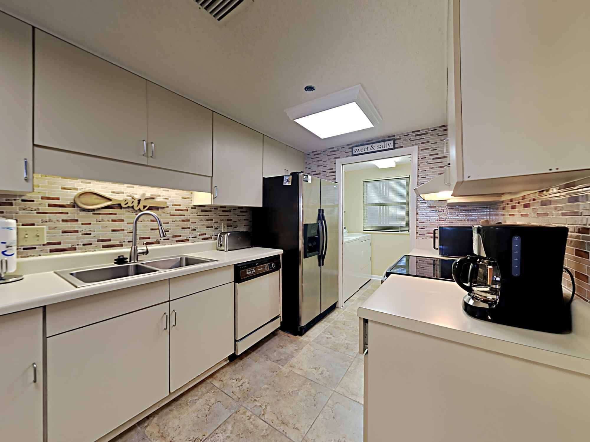 A modern kitchen with white cabinets, a dishwasher, a refrigerator, a coffee maker, and a window with blinds.
