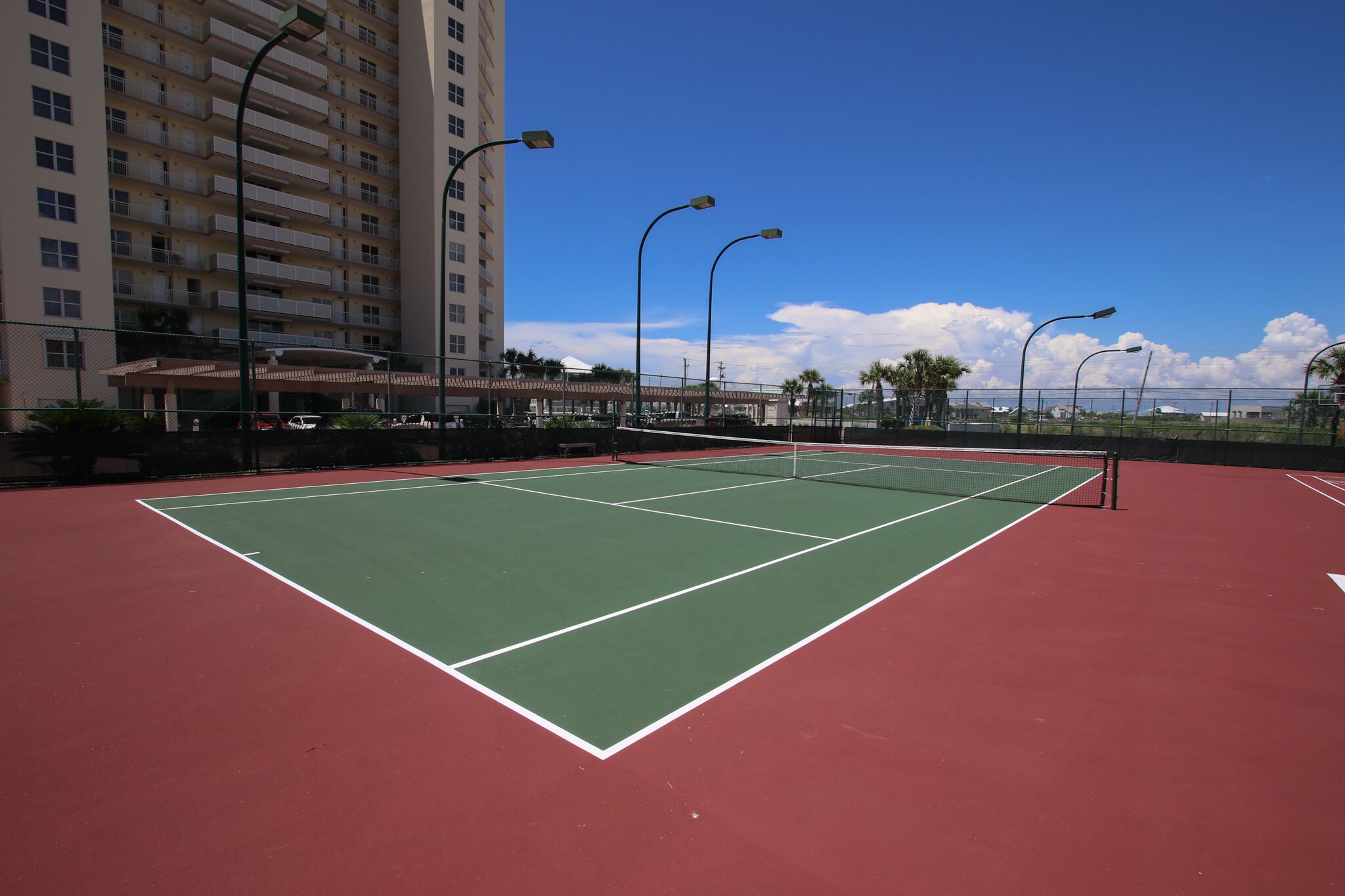 A tennis court with a net, surrounded by a fence, and a tall building in the background.