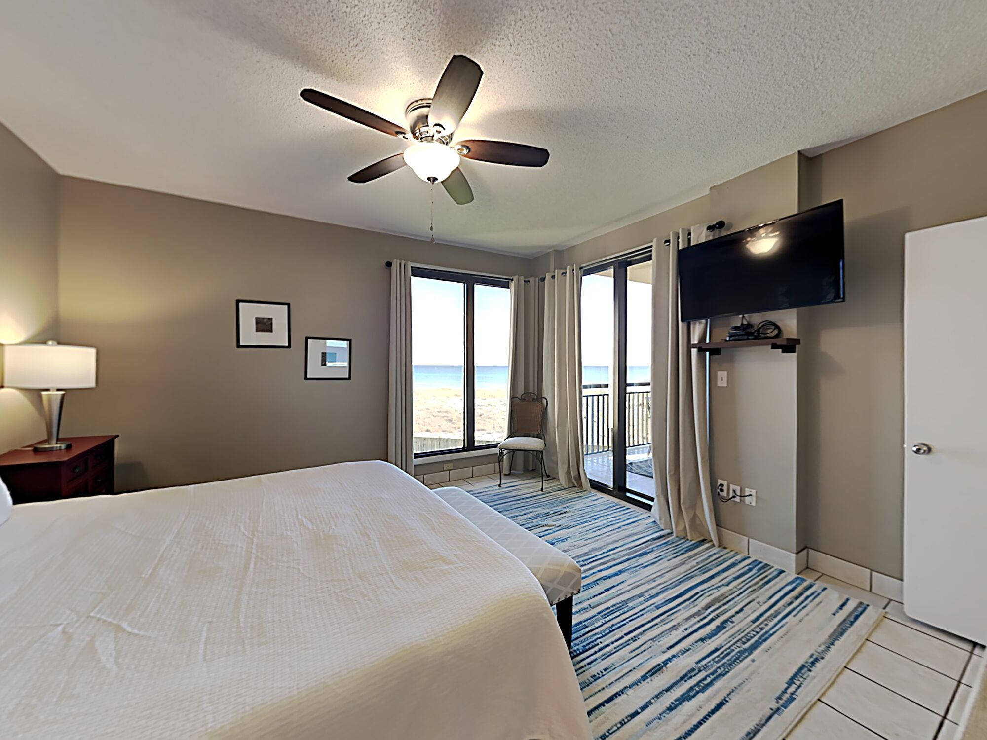 Habitación de hotel con cama, ventilador de techo, televisor, silla y vista a la playa a través de las puertas del balcón.