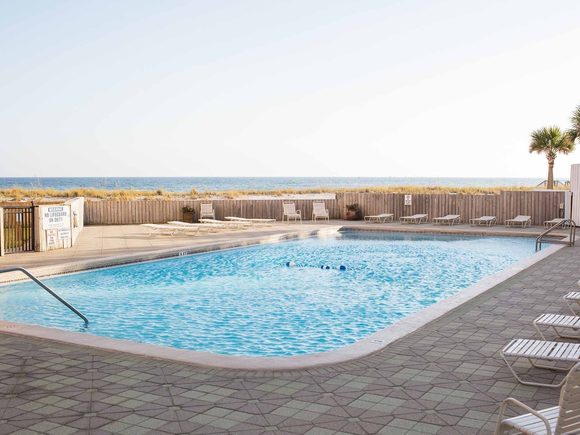 A swimming pool with lounge chairs and a wooden fence, with a beach view in the background.