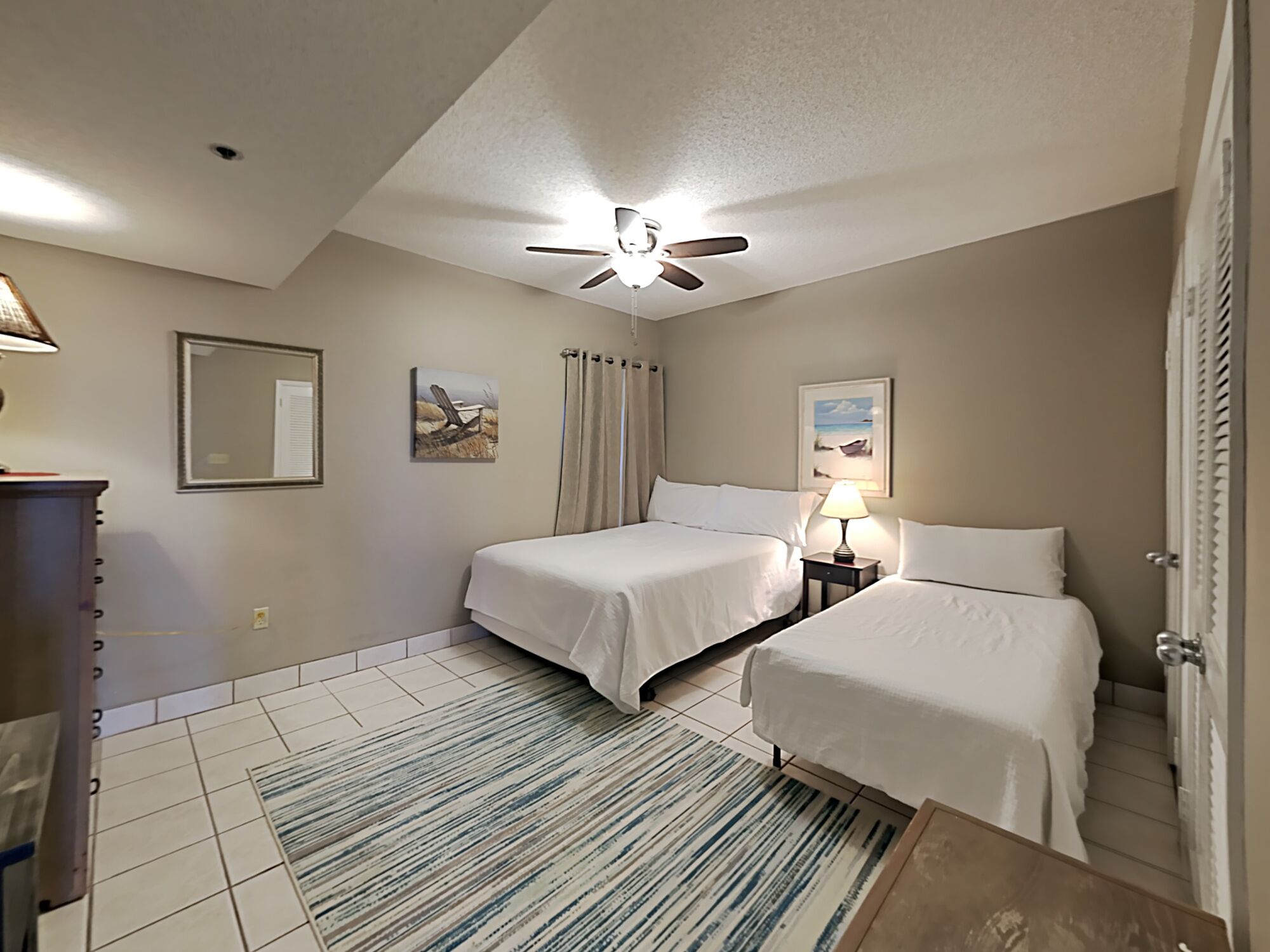 Quarto de hotel com duas camas, um ventilador de teto, uma mesinha com abajur e uma janela com cortinas.