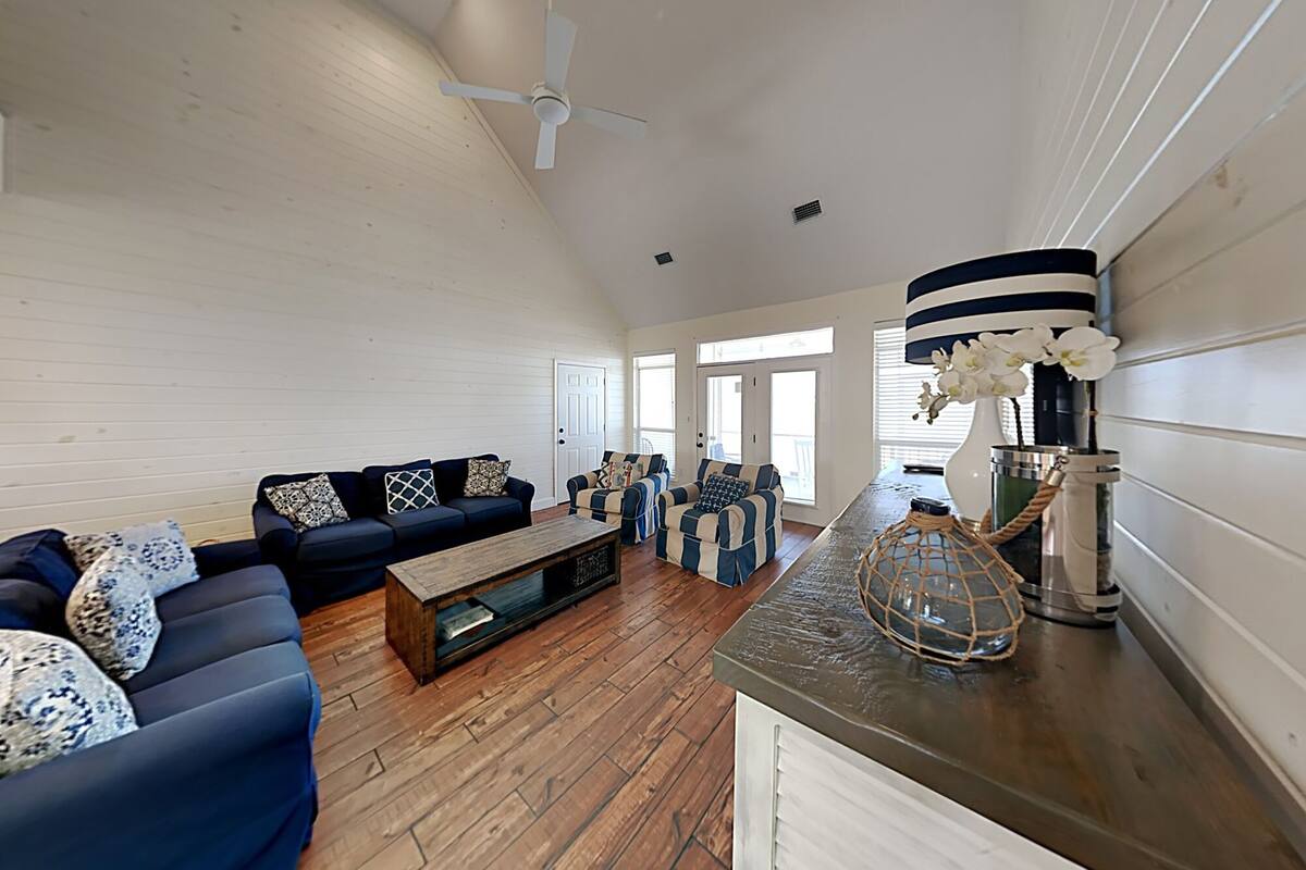 A spacious living room with a dark blue sofa set, a wooden coffee table, and a striped armchair. A ceiling fan is visible, and the room has wooden flooring.