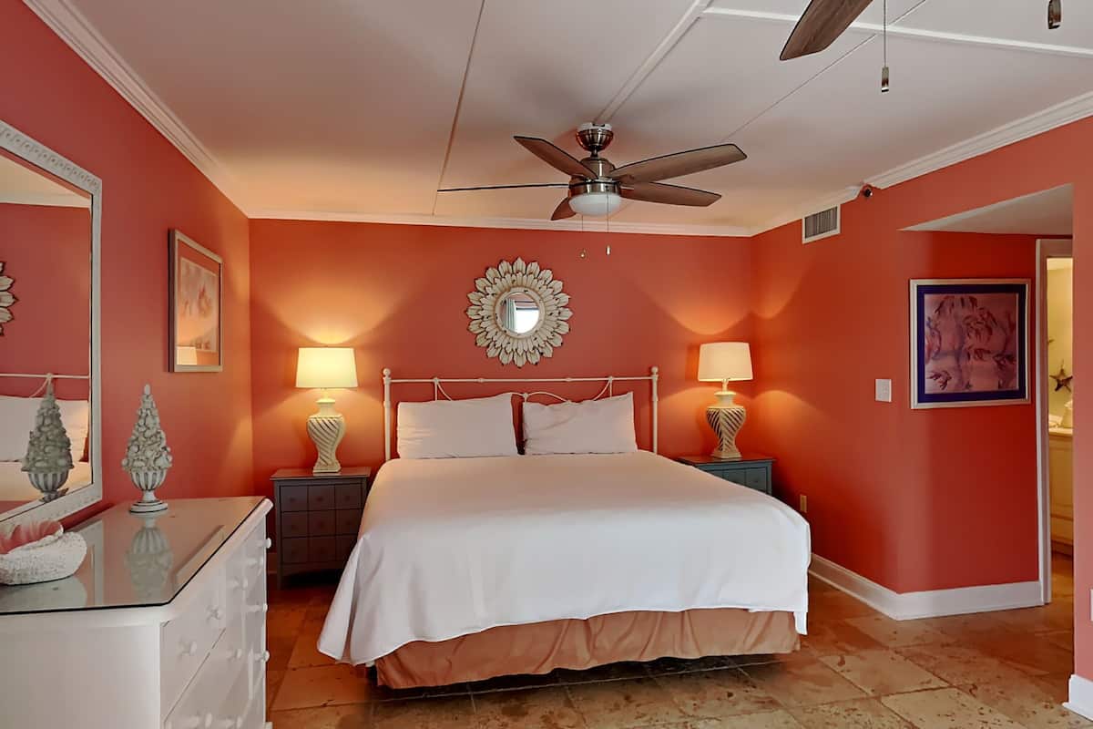 A bedroom with a white bed, two nightstands with lamps, a ceiling fan, and a large mirror on the wall.