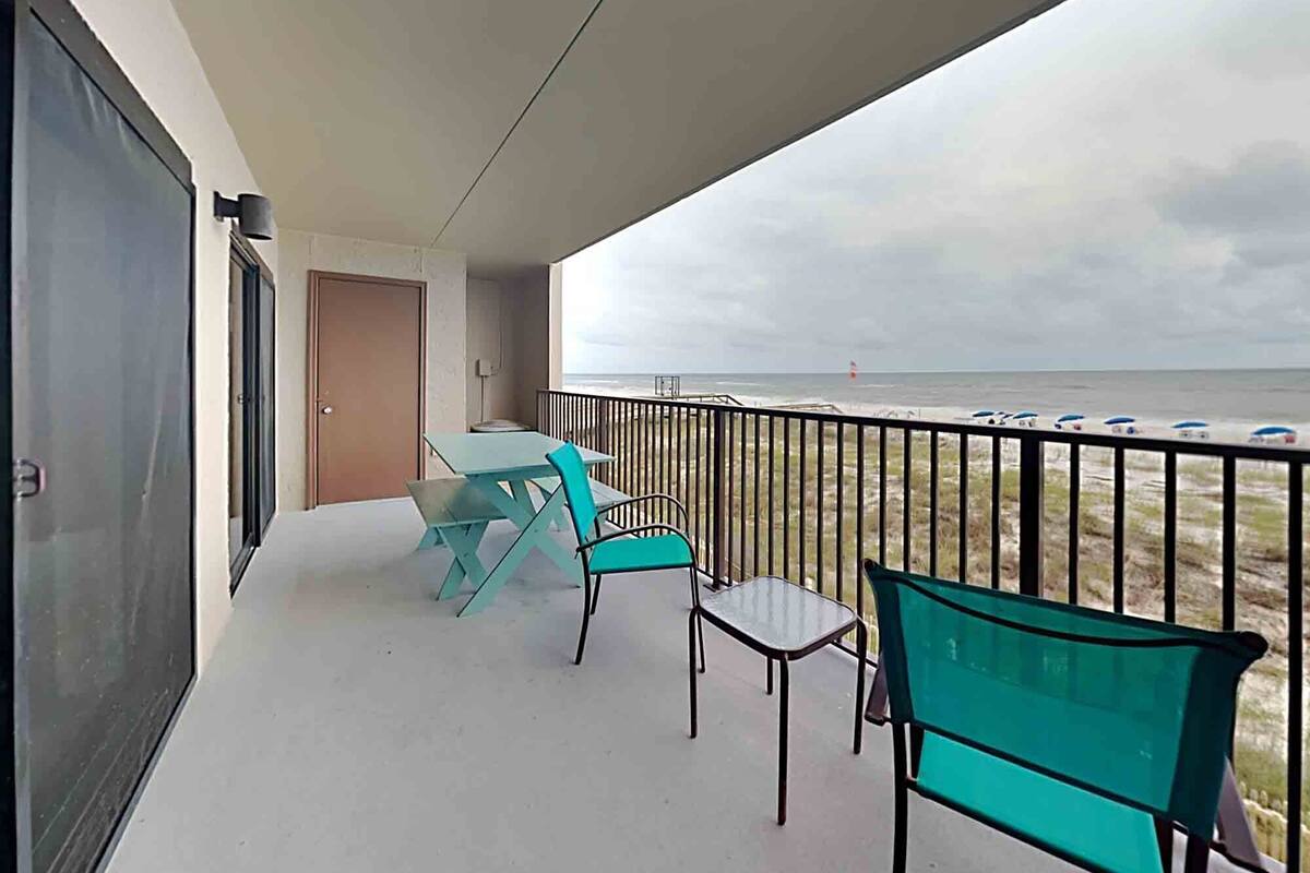 A balcony with turquoise chairs and a table, overlooking a beach with umbrellas.