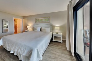 A spacious bedroom with a large bed, bedside tables with lamps, a door leading to another room, and a window with curtains. - Ocean Breeze West by Southern Vacation Rentals (Pensacola)