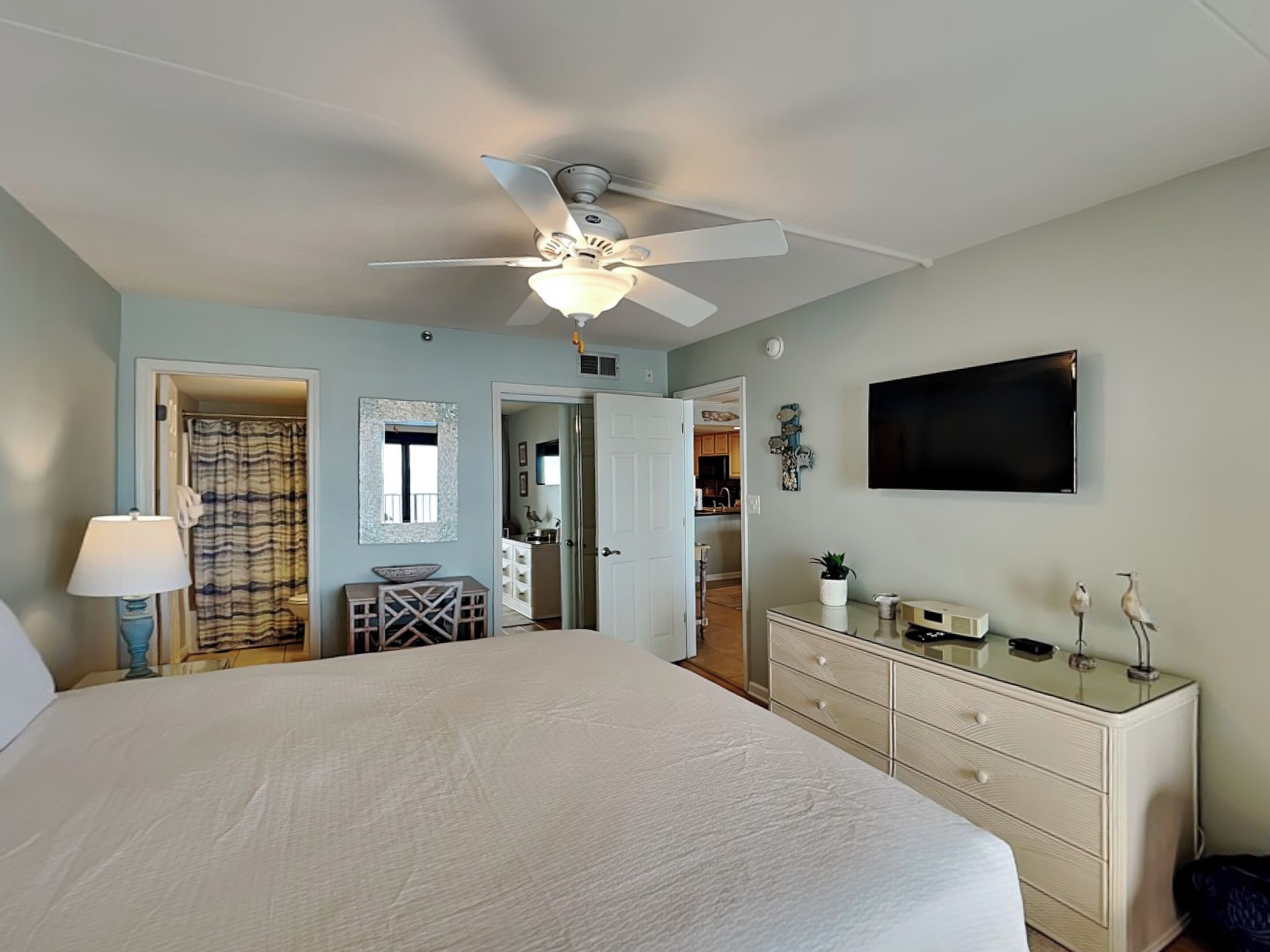A bedroom with a bed, a ceiling fan, a dresser, a TV, and a bathroom visible through an open door.