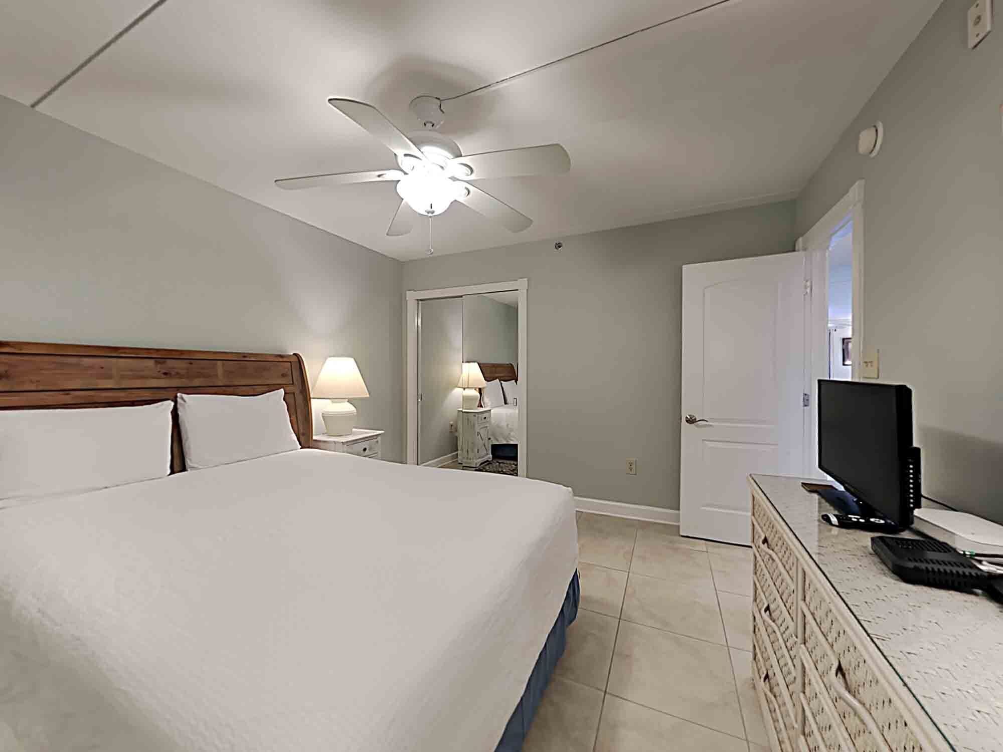A hotel room with a bed, a desk with a computer, a television, a ceiling fan, and a door.
