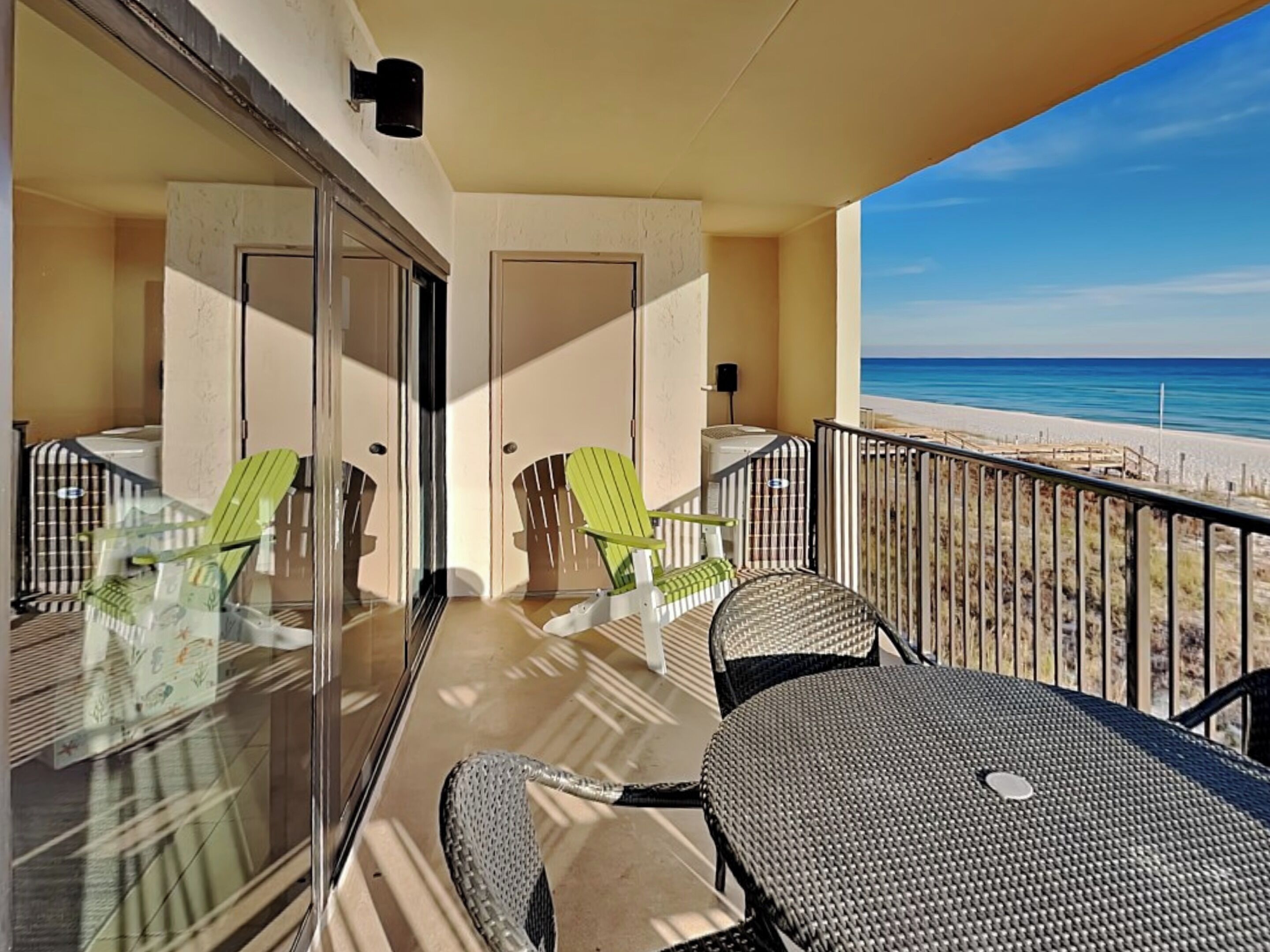 Uma varanda com porta de vidro, duas cadeiras verdes, uma mesa de vime e vista para a praia.