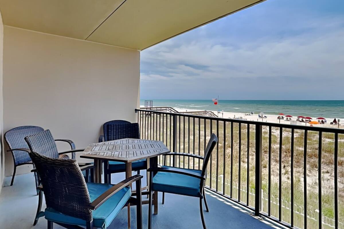 A balcony with a table and chairs, overlooking a beach with umbrellas and a flag.