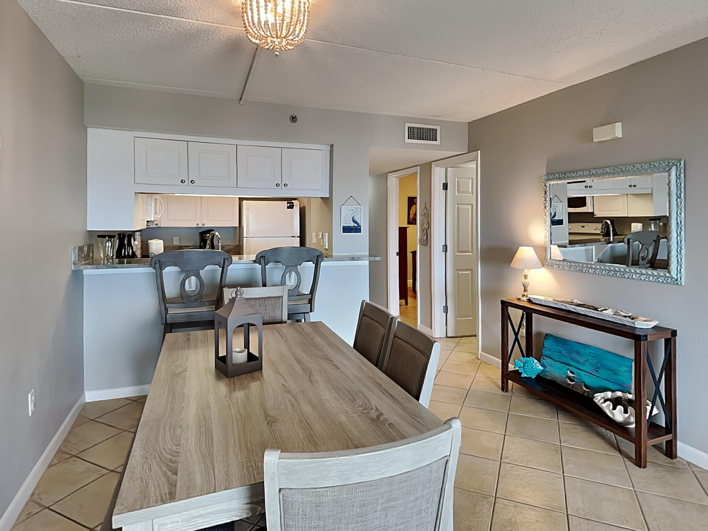 A dining area with a wooden table and chairs, a kitchen in the background, and a mirror on the wall.