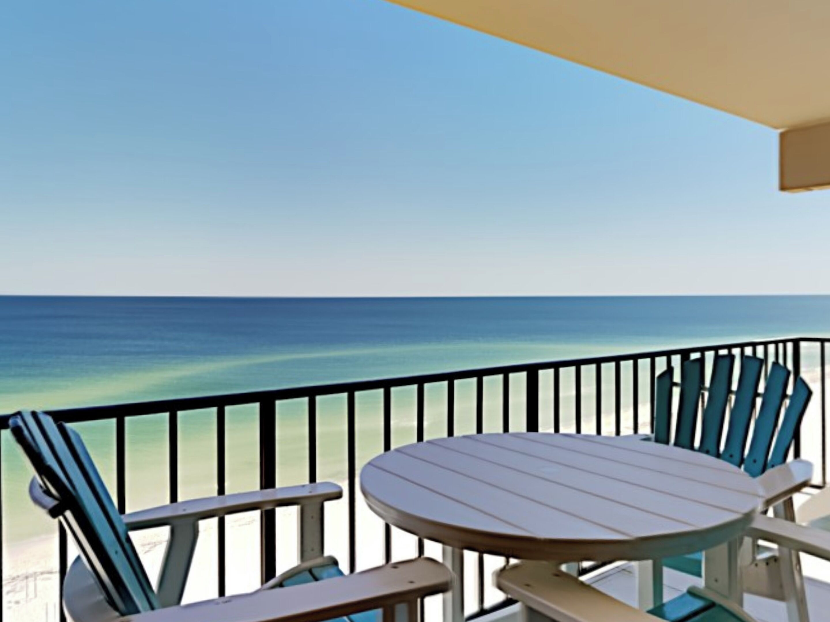 Ein Balkon mit Tisch und Stühlen, mit Blick auf einen Strand und klares, blaues Wasser.
