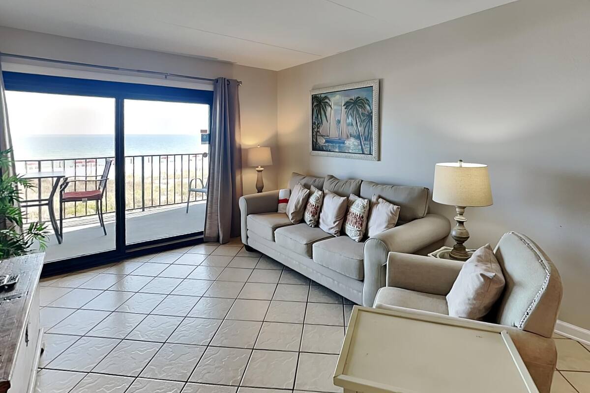 A living room with a sofa, a small table, a lamp, a painting, and a view of the ocean through a sliding door.