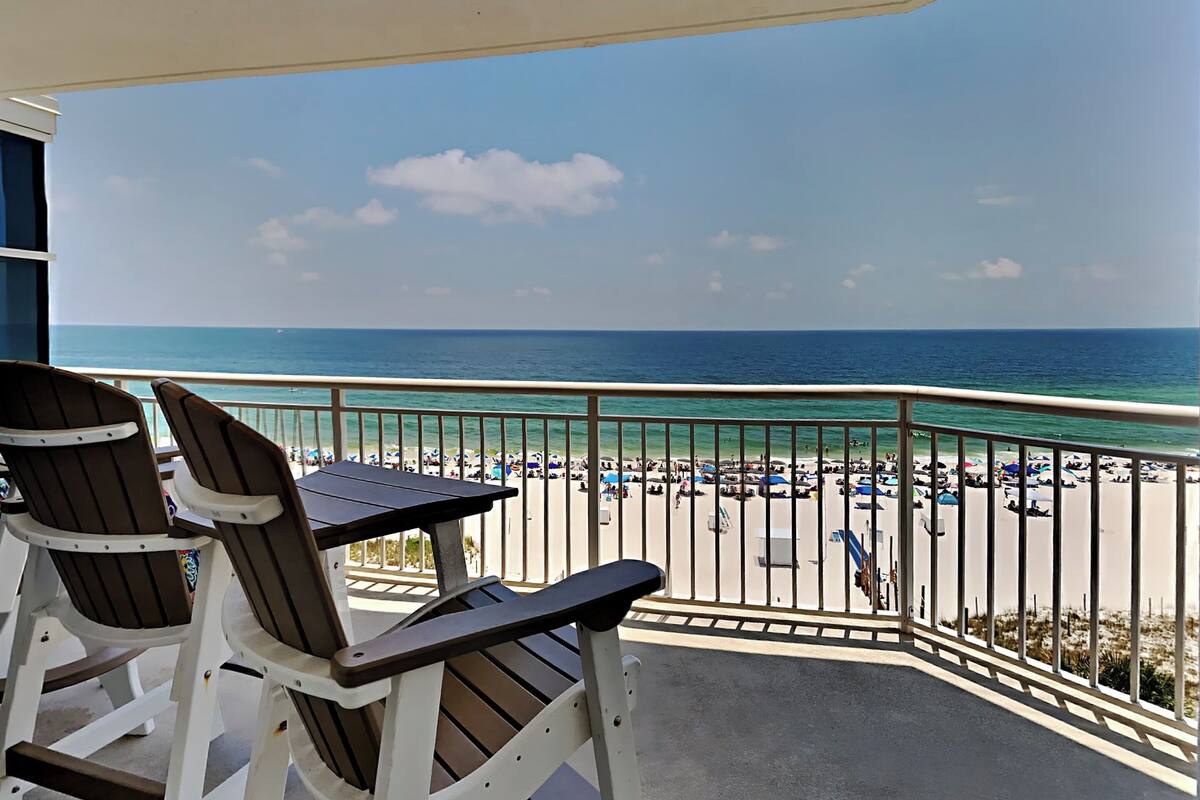 A balcony with two chairs and a table, overlooking a beach with umbrellas and people.