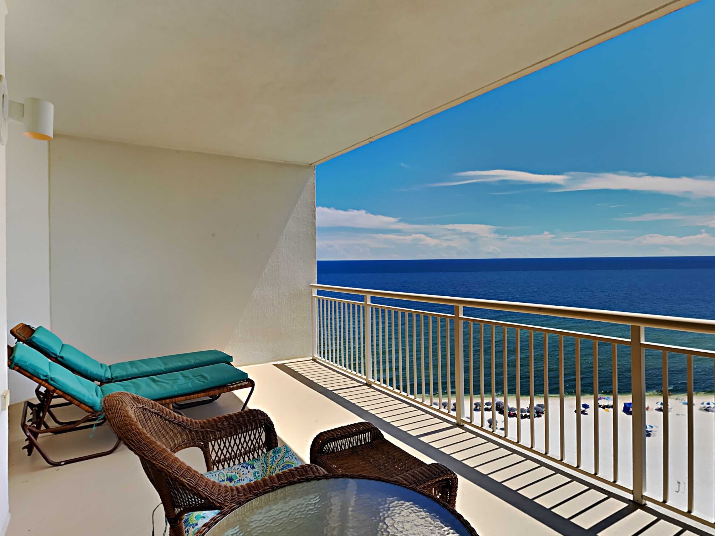 Een balkon met rieten meubels en een glazen tafel, met uitzicht op een strand en een helderblauwe lucht en zee.