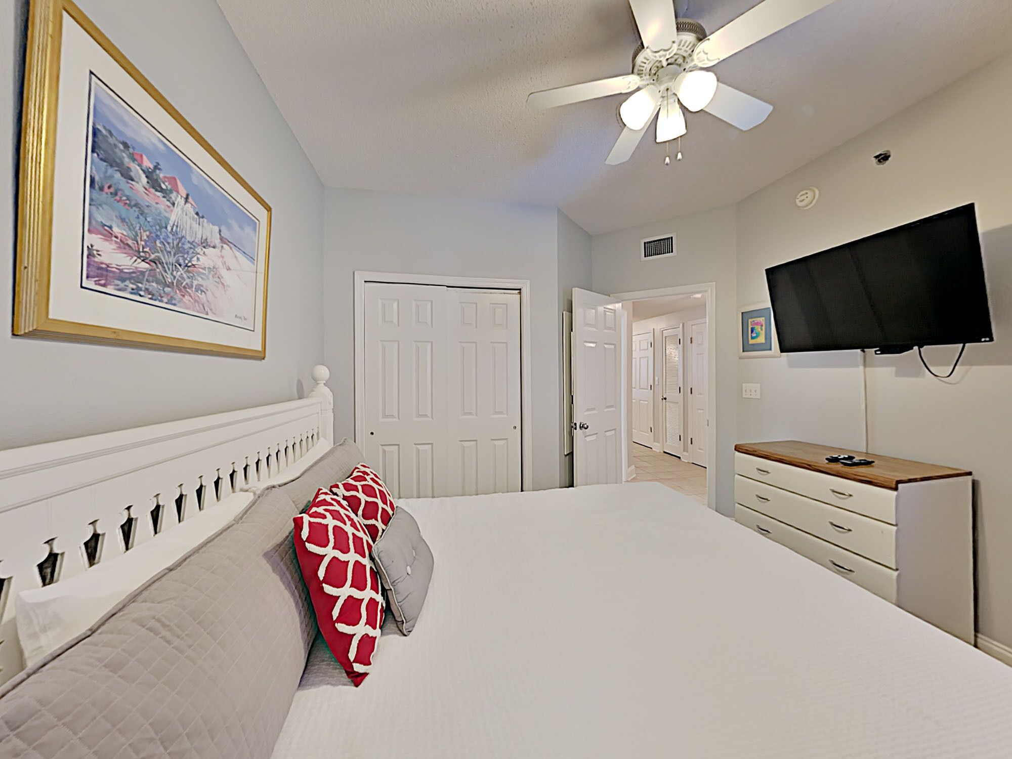 A bedroom with a bed, a ceiling fan, a TV, and a door leading to another room.
