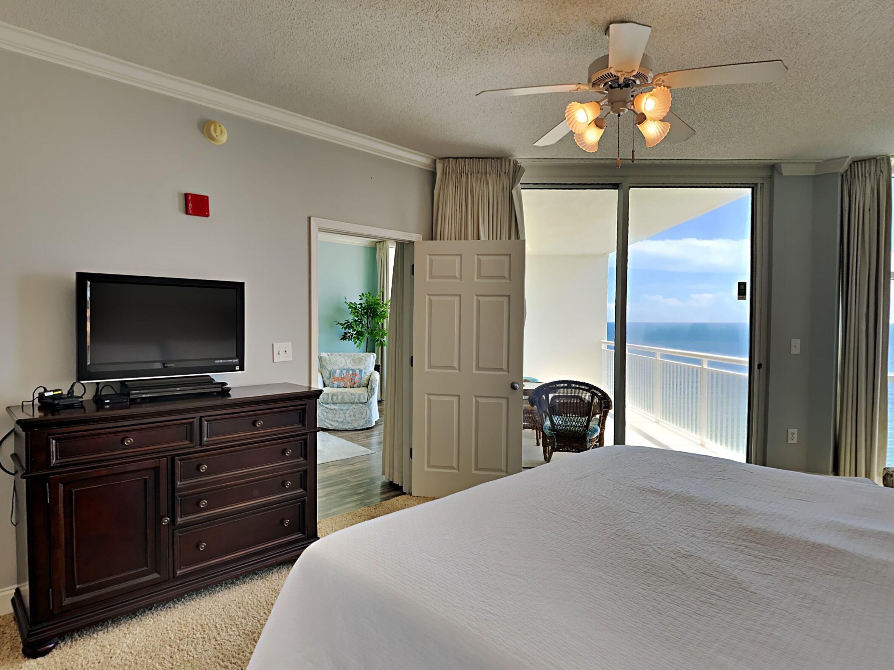 A hotel room with a large bed, a TV on a dresser, a ceiling fan, and a balcony with a view of the ocean.