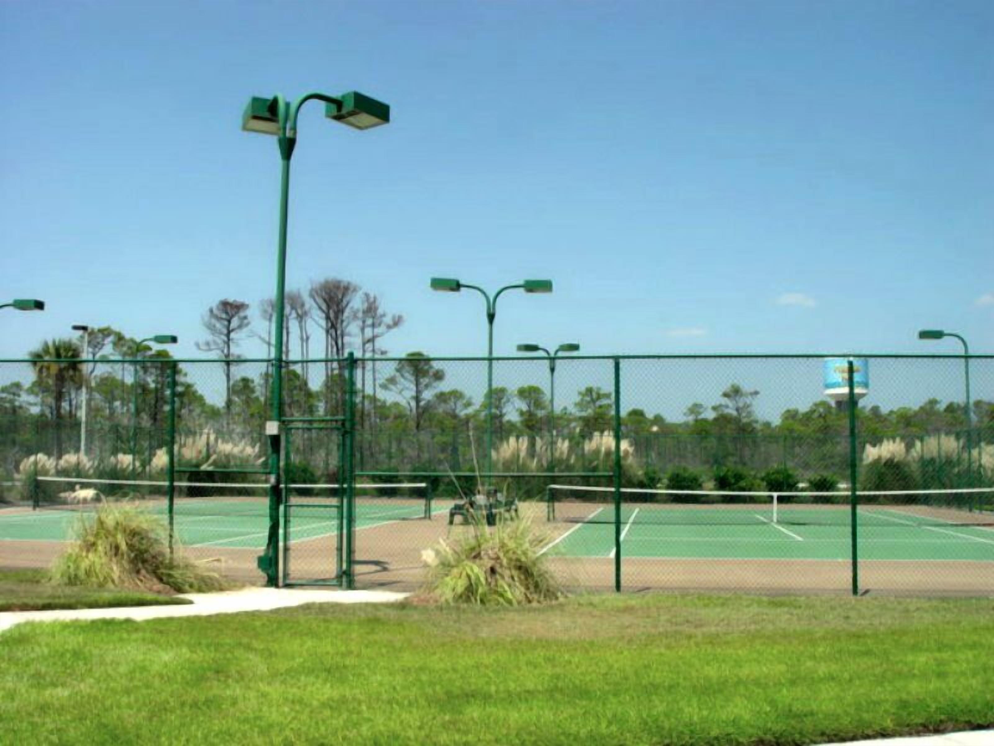 A tennis court with green fencing, street lamps, and a clear sky.