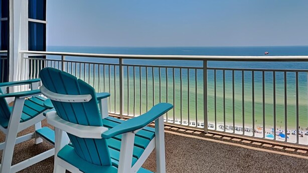 A balcony with two teal Adirondack chairs, a clear view of the ocean, and a sandy beach with umbrellas in the distance.