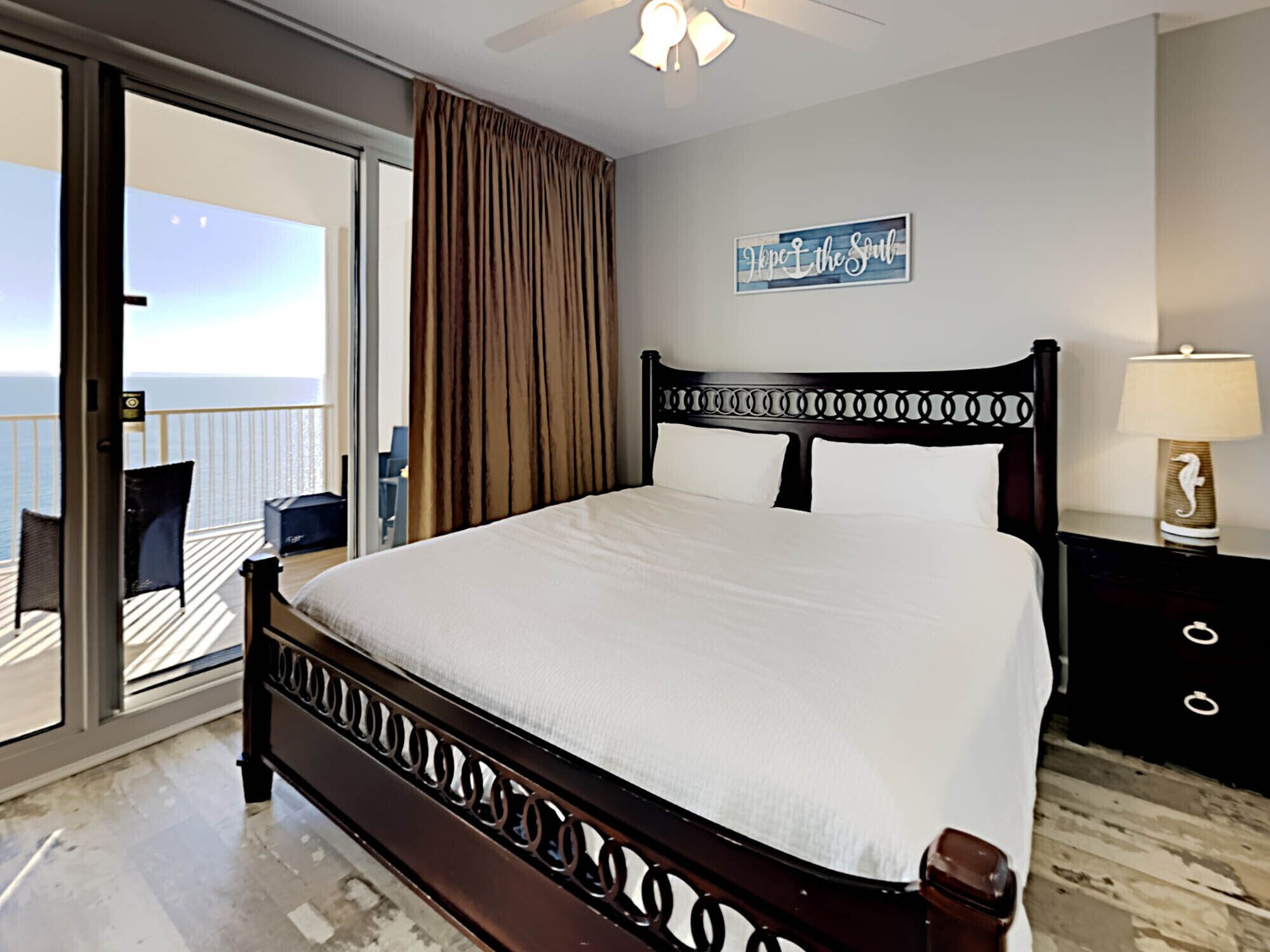 A bedroom with a large bed, a view of the ocean, a ceiling fan, and a sign on the wall.