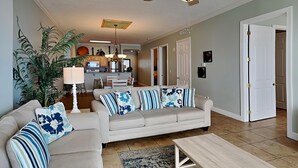 A living room with a beige sofa, a coffee table, and a ceiling fan. There is a kitchen in the background with a dining area. - Ocean Reef by Resort Collection (Panama City Beach)
