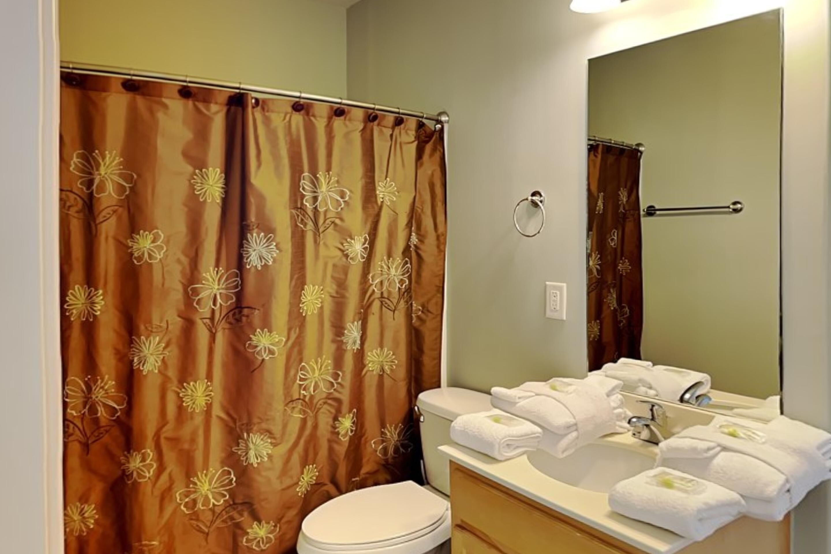 A bathroom with a shower curtain featuring a floral pattern, a toilet, a sink with multiple towels, and a mirror.
