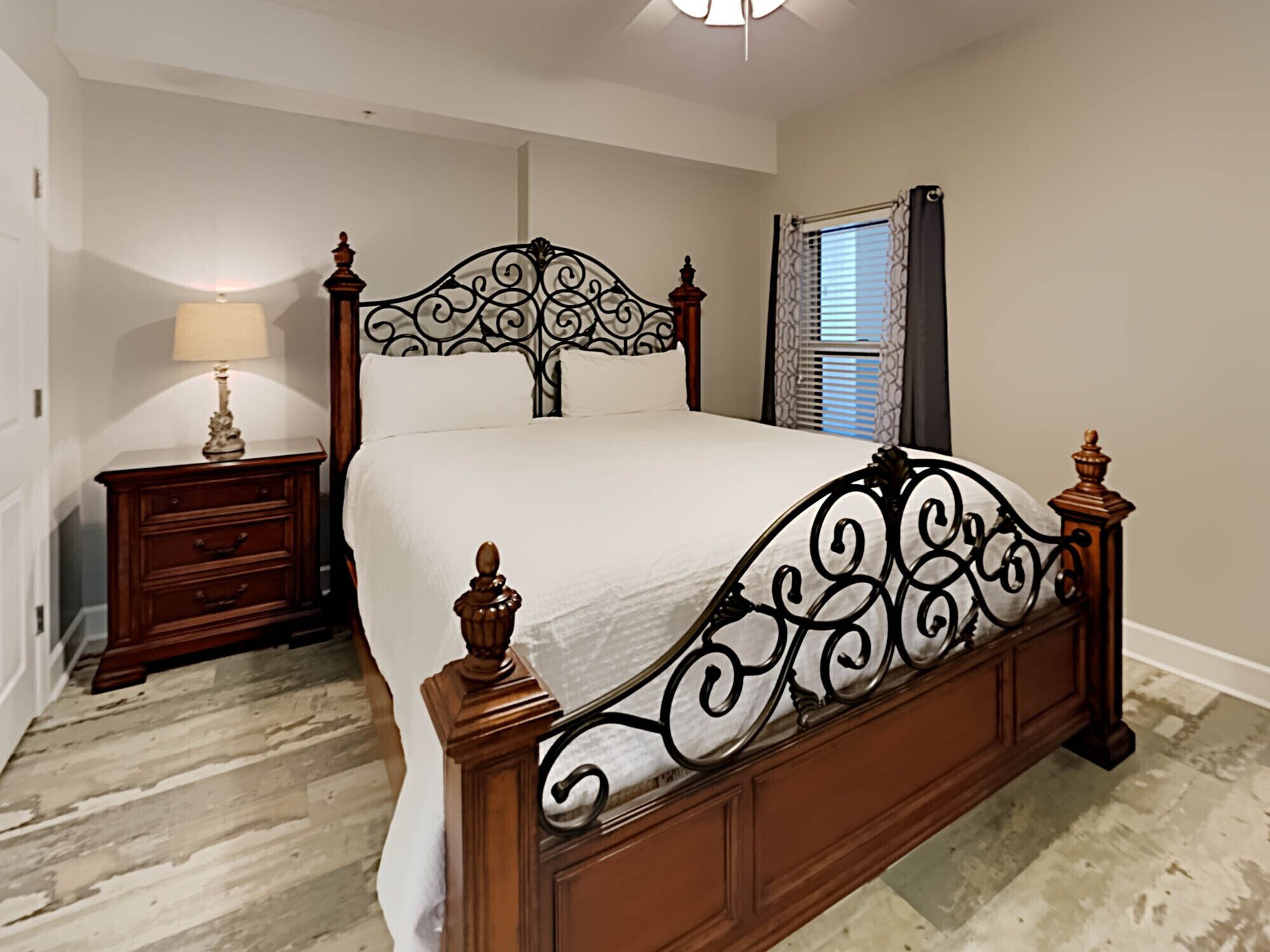 A bedroom with a wooden bed frame, a nightstand with a lamp, and a window with blinds.