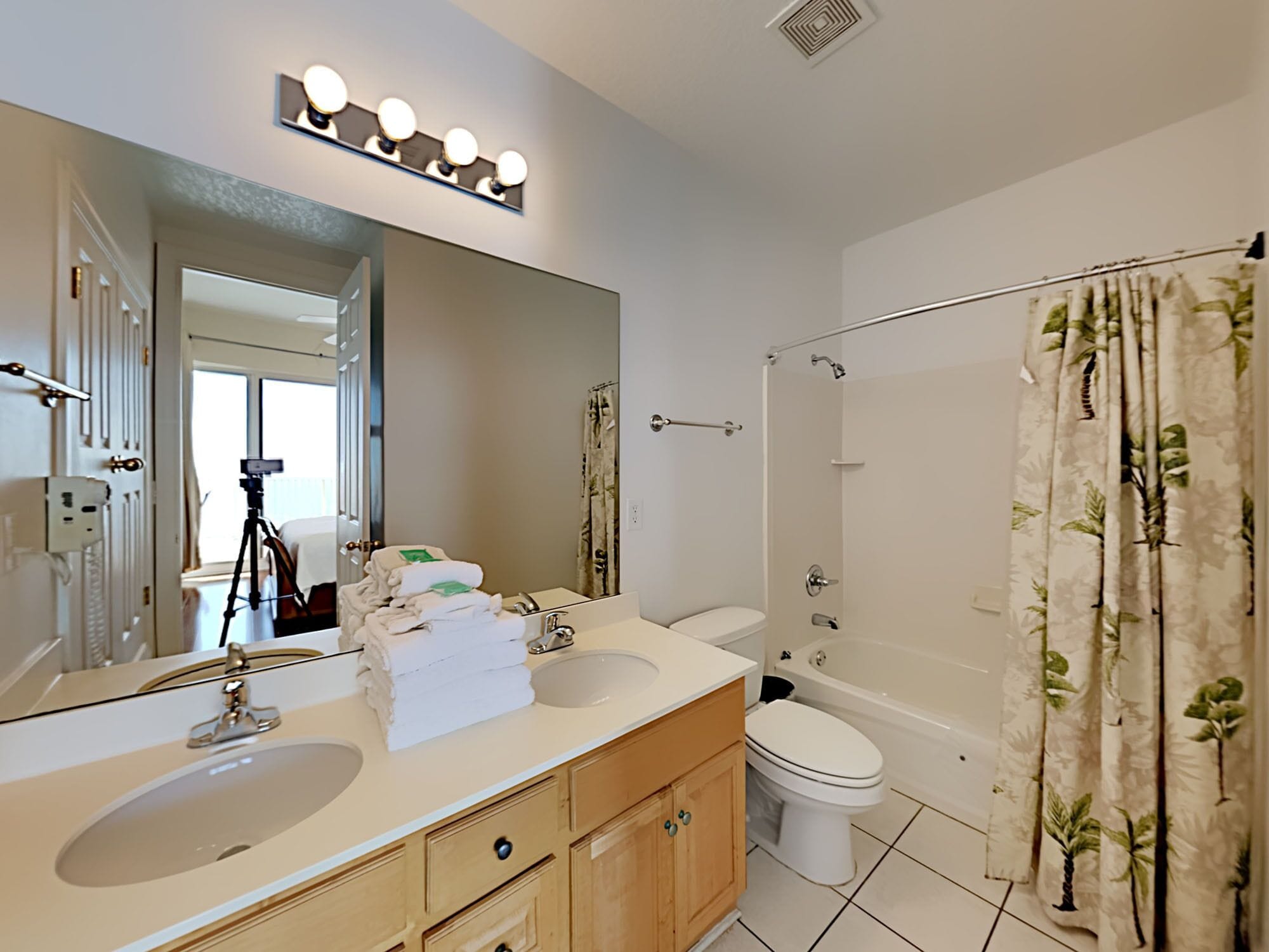 Une salle de bain avec deux lavabos, des toilettes, une baignoire et une douche. On y trouve un miroir avec des lumières au-dessus, et un trépied pour appareil photo est visible.