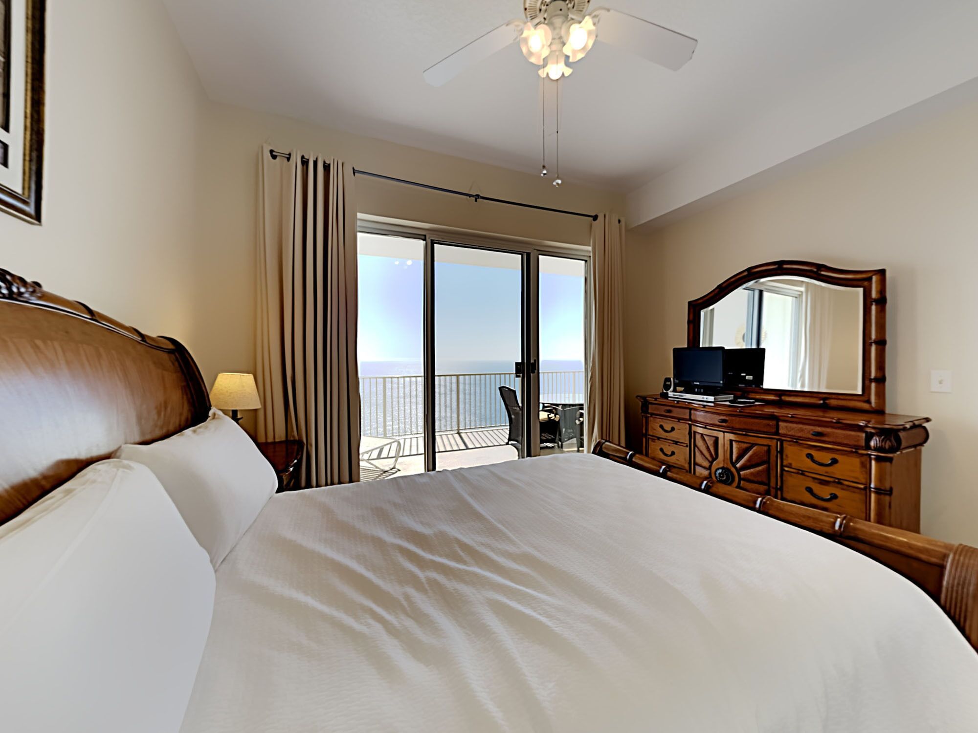 A hotel room with a large bed, a dresser with a mirror, a desk with a chair, and a balcony with a view of the ocean.