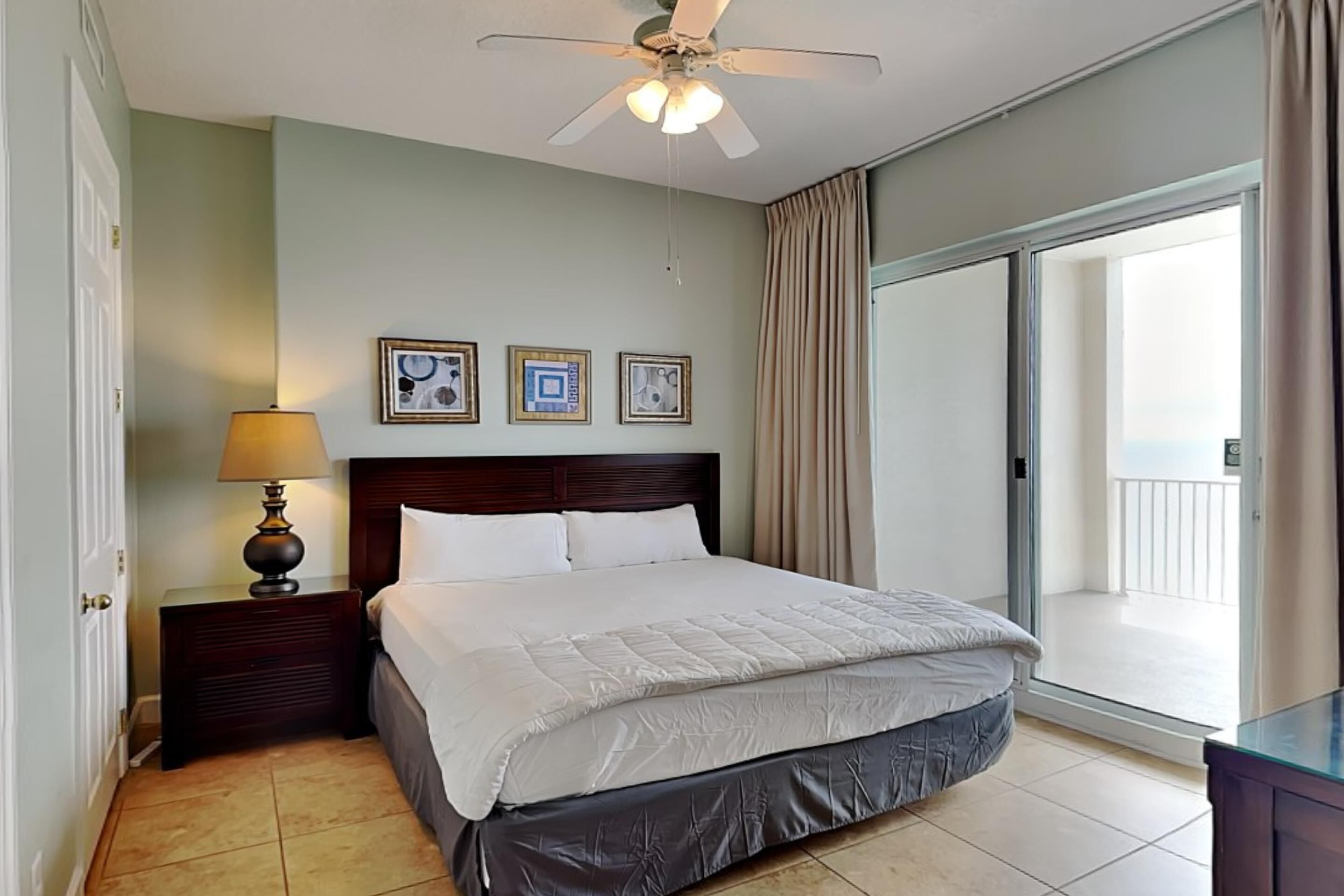 Une chambre d’hôtel avec un lit, une table de chevet avec une lampe, un ventilateur de plafond et une vue sur le balcon extérieur.