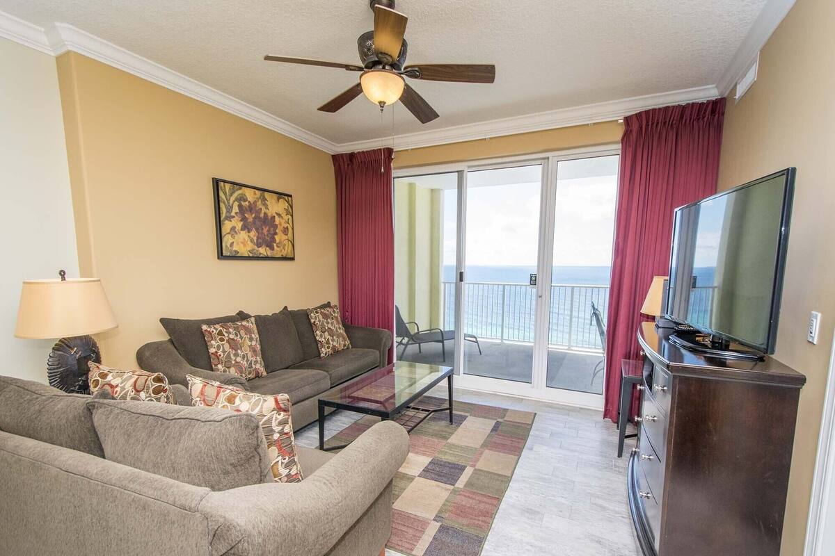 Een woonkamer met een bank, fauteuil, salontafel en een televisie. Er is een balkon met uitzicht op de oceaan.