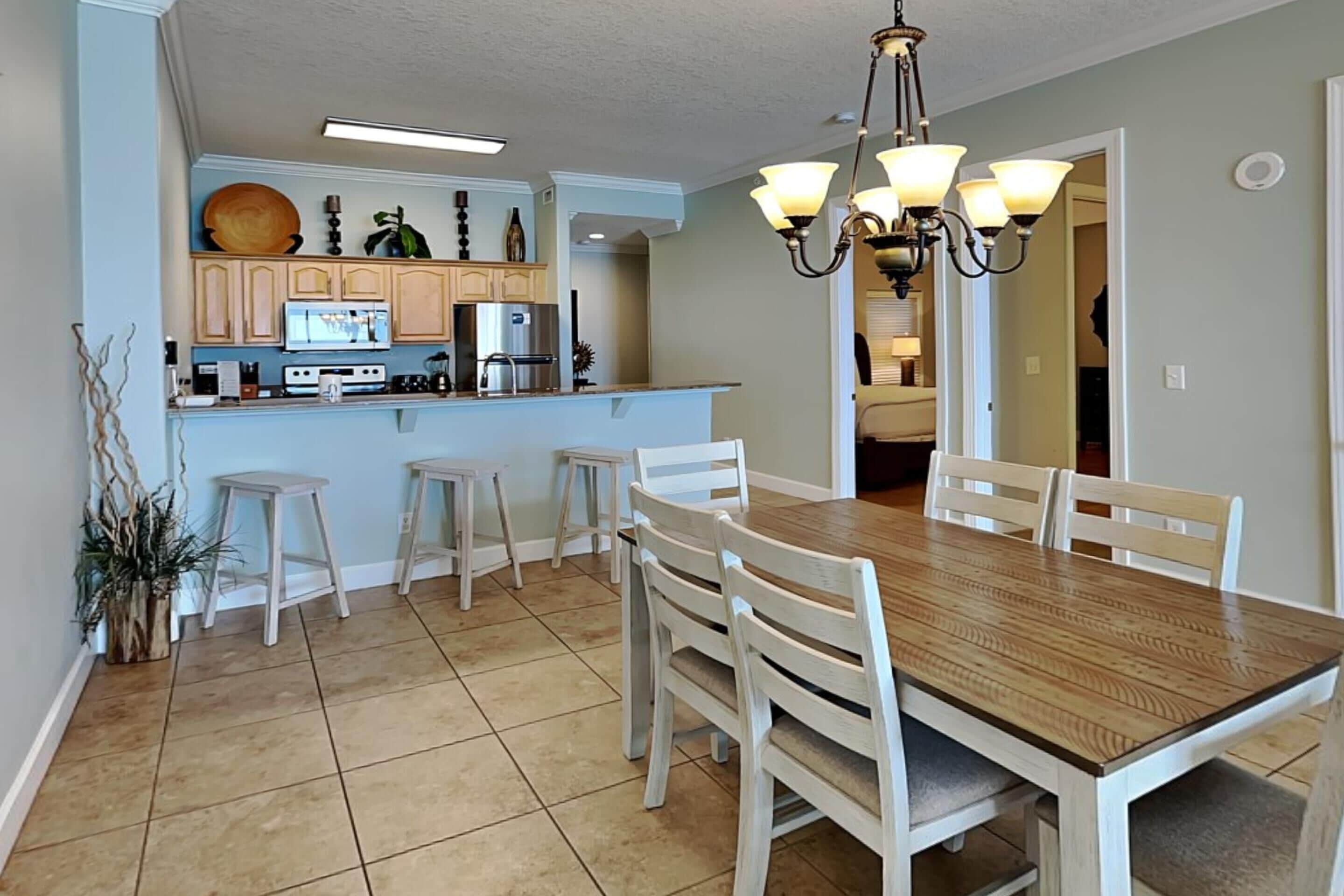 A dining area with a wooden table and white chairs, a kitchen with light-colored cabinets, and a chandelier.