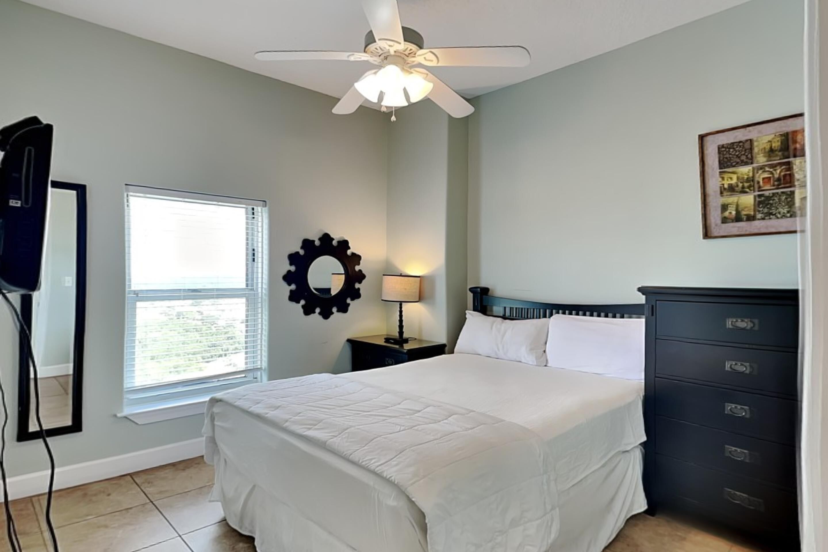 A bedroom with a bed, a nightstand, a dresser, a ceiling fan, a window with blinds, and a wall-mounted mirror.