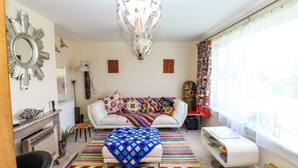 A living room with a white sofa, colorful throw pillows, a patterned rug, a chandelier, and a large window with curtains. - Jasper's Corner (Kingsbridge)