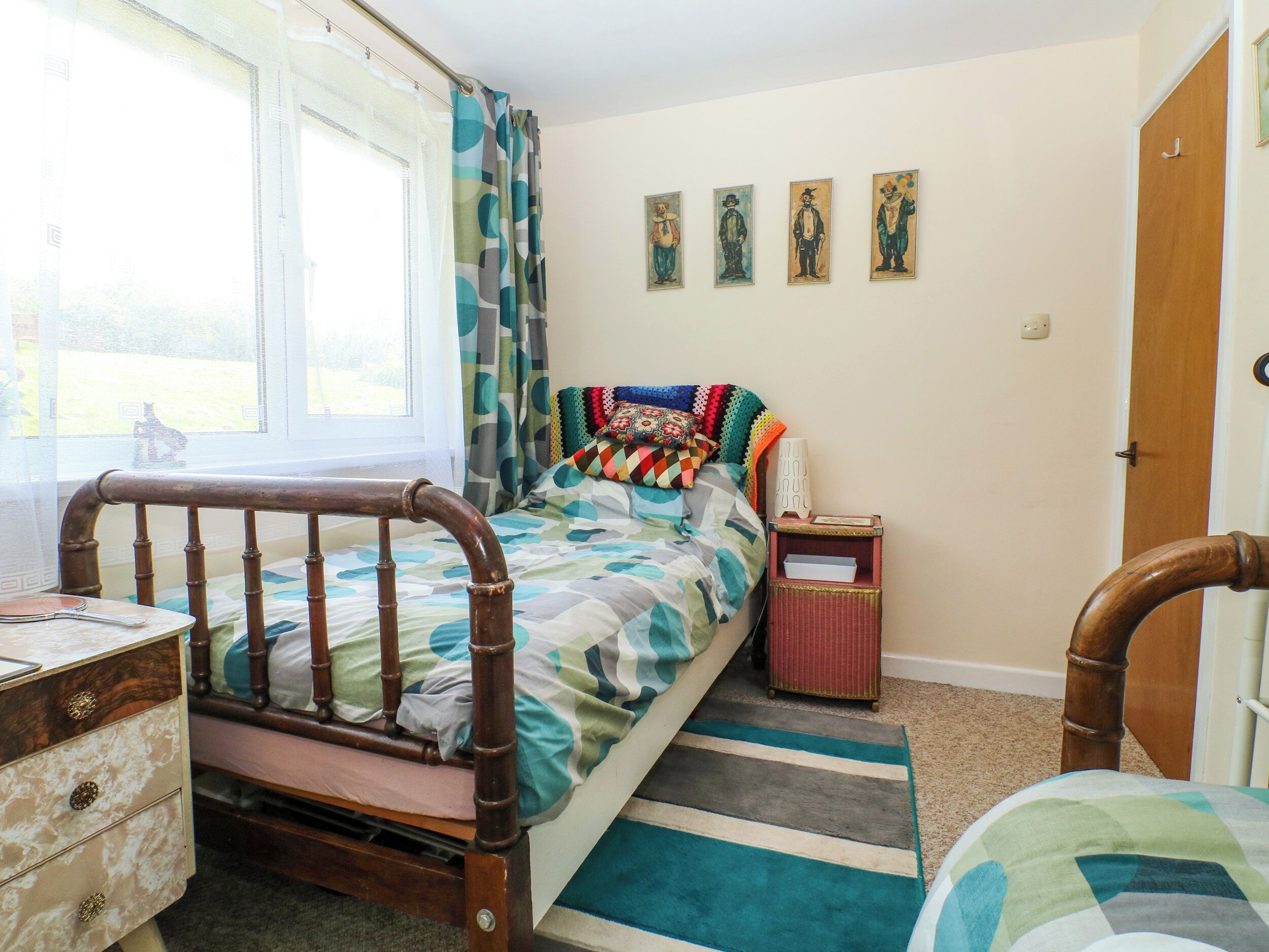 Une chambre à coucher avec un lit en bois, une table de chevet, une commode, une fenêtre avec des rideaux et un mur orné de tableaux encadrés.