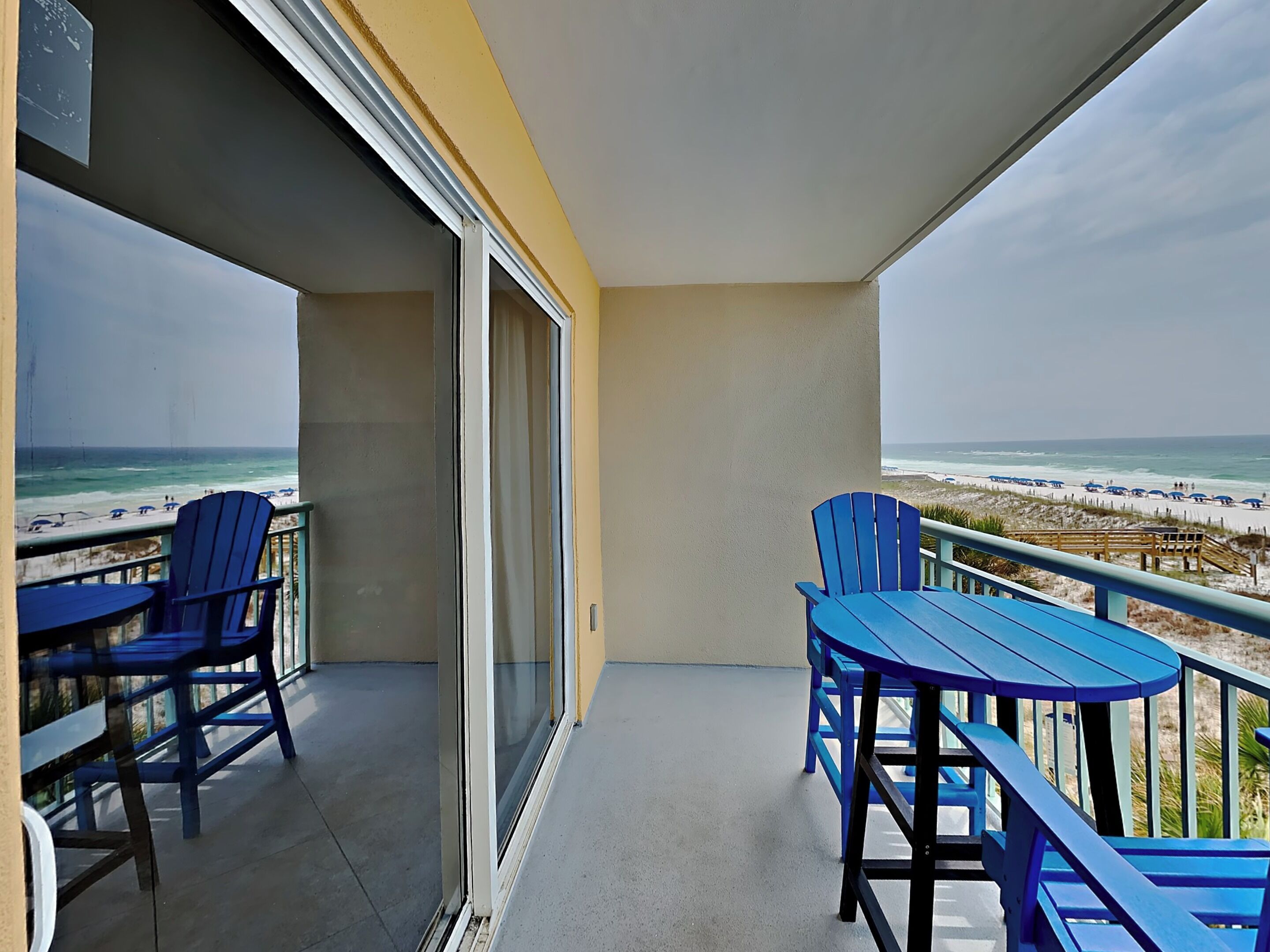 A balcony with blue chairs and a table, overlooking a beach with umbrellas.