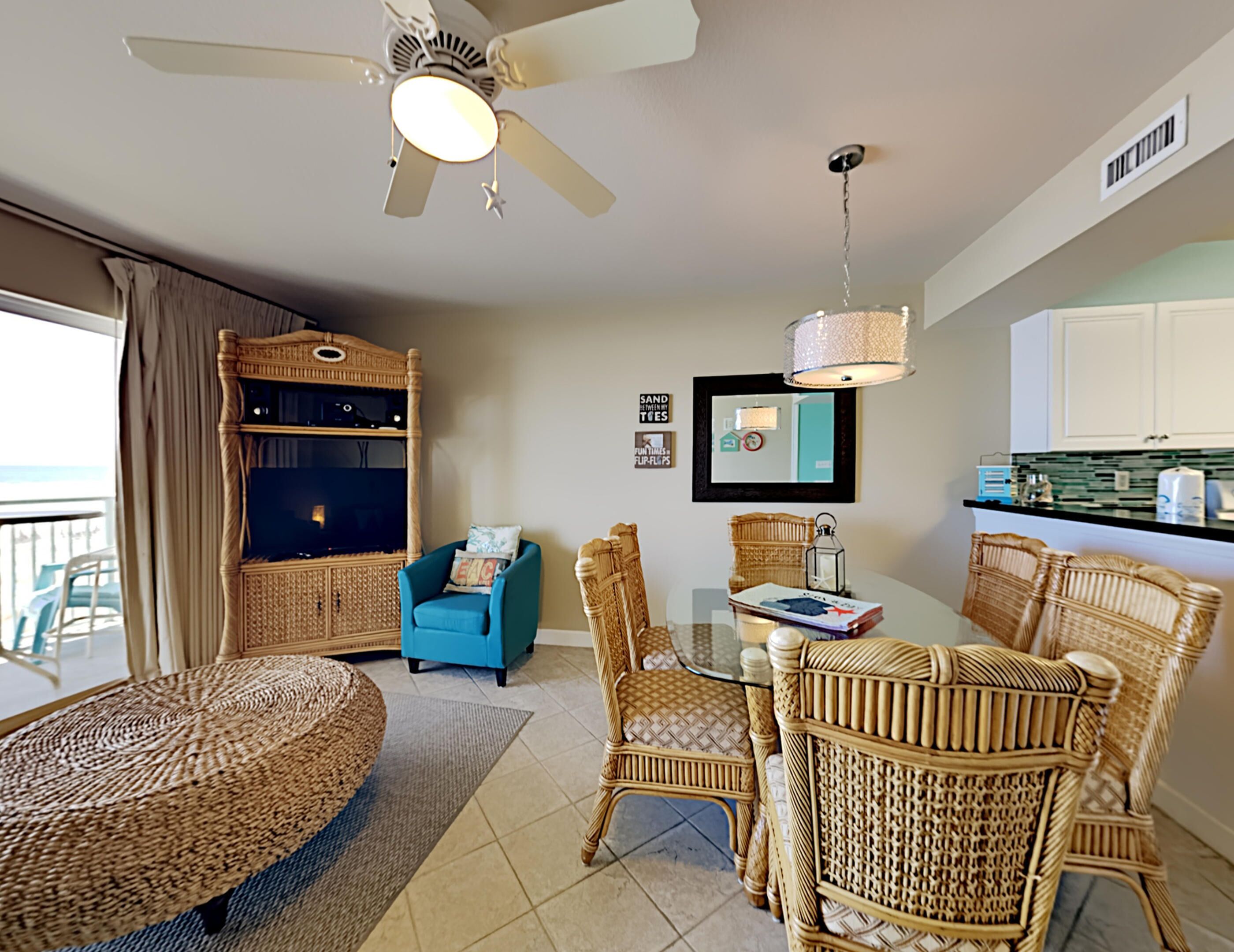 A living room with a ceiling fan, wicker furniture, a blue armchair, a glass table, and a kitchen area with white cabinets.