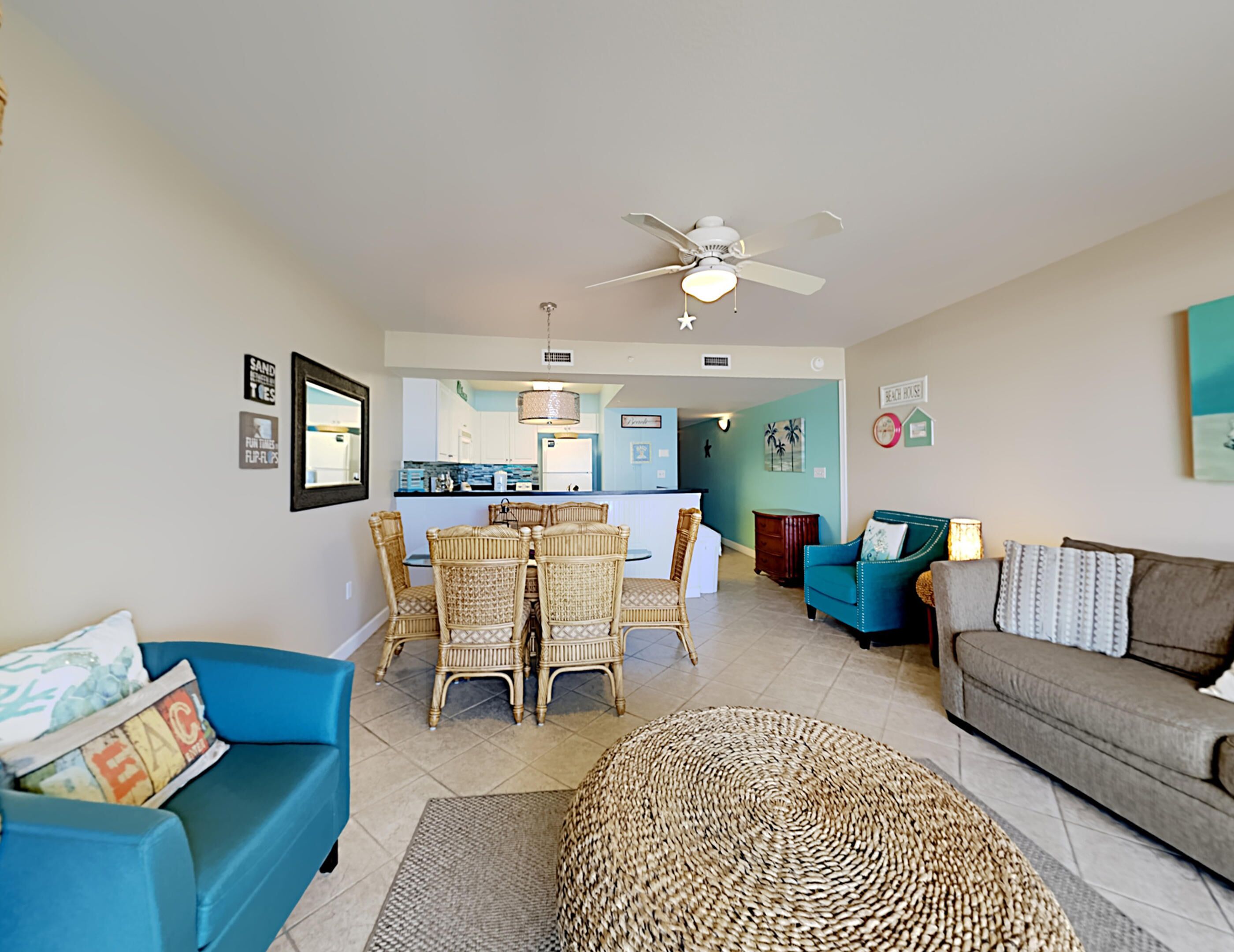 A living room with a blue sofa, a grey sofa, wicker chairs, a woven rug, and a ceiling fan.