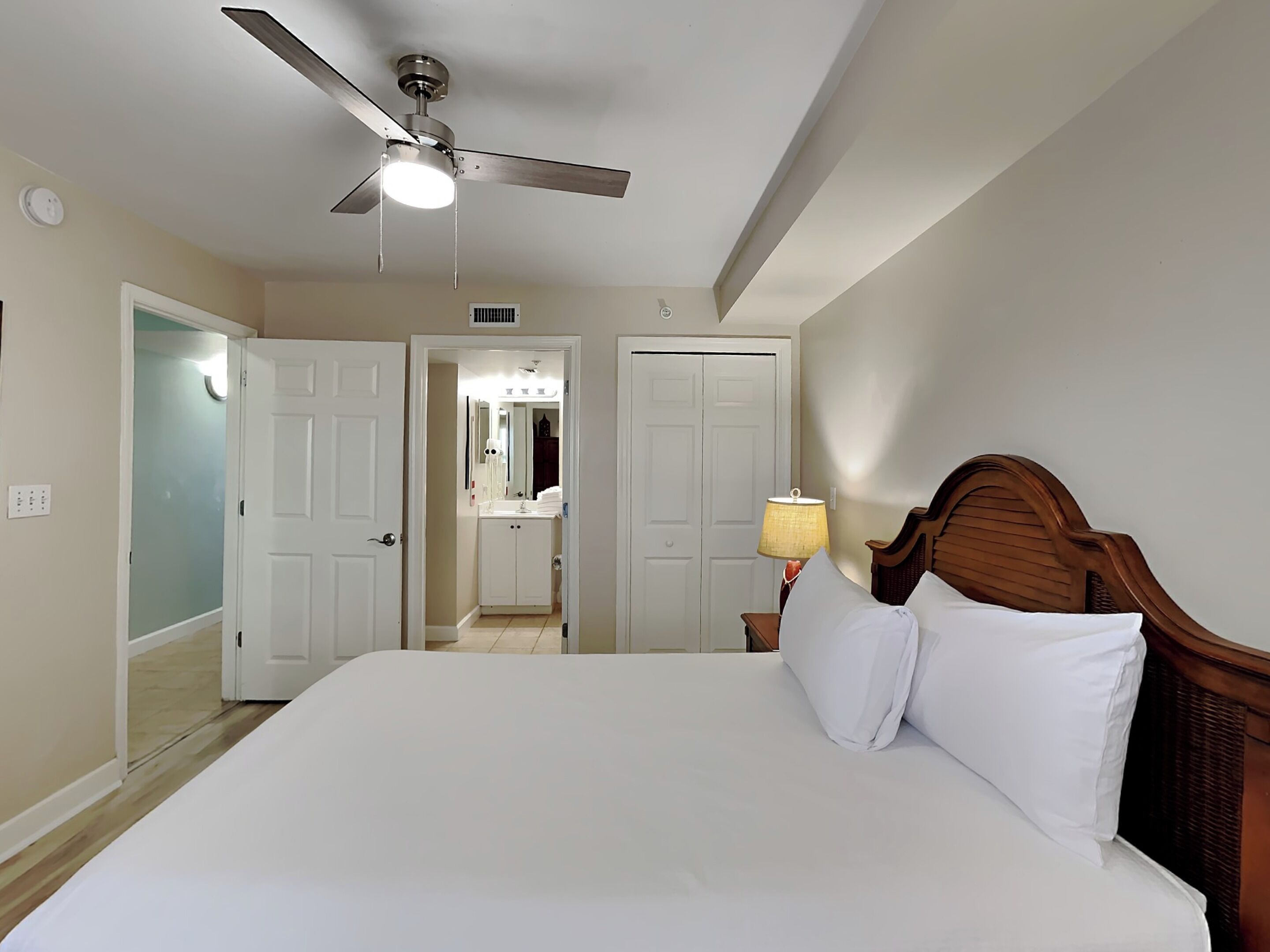A hotel room with a bed, a ceiling fan, a nightstand with a lamp, and a bathroom visible through an open door.