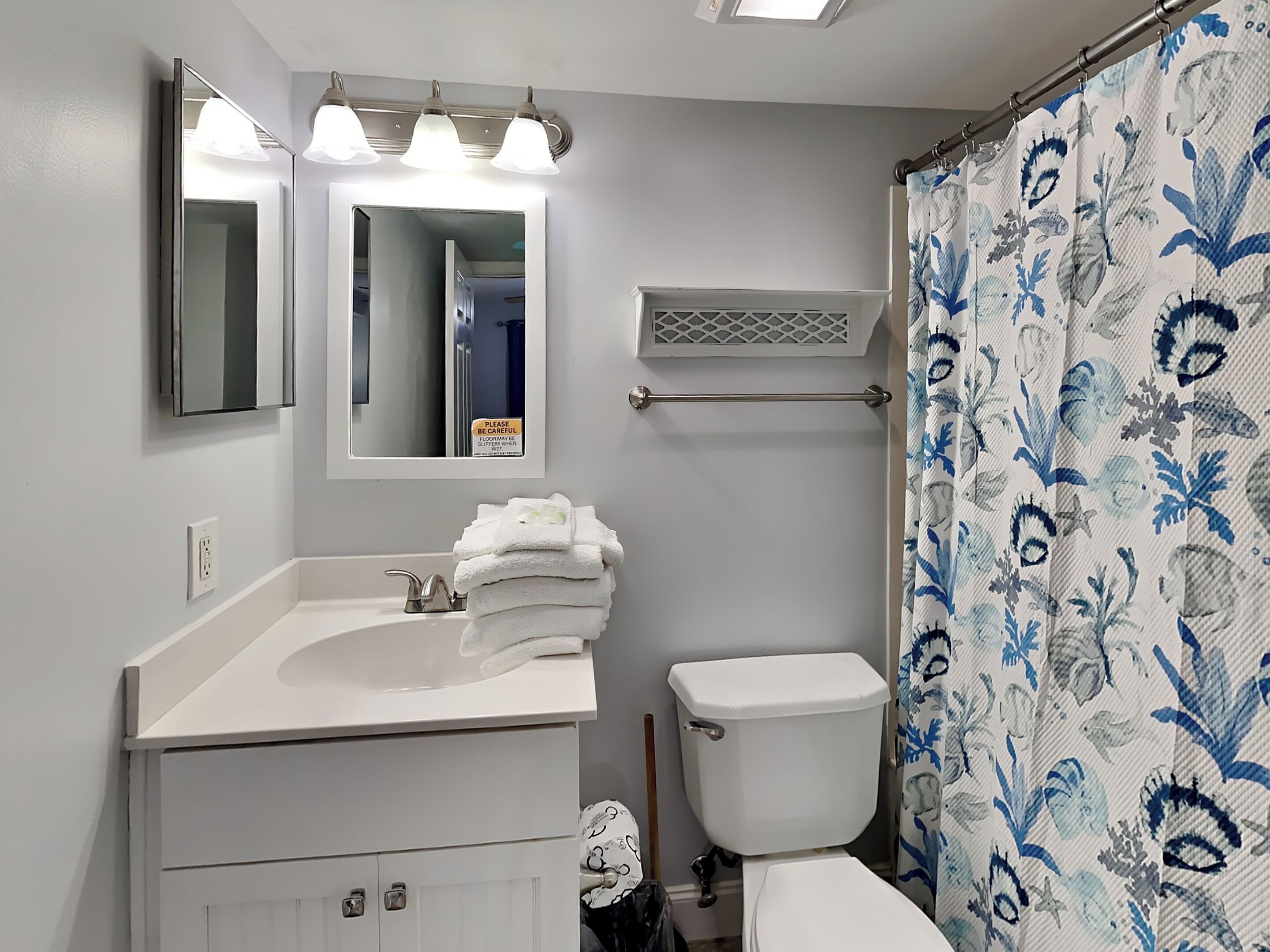 Un baño con un lavamanos blanco, espejo e inodoro. Hay un perchero con toallas dobladas y una cortina de ducha con diseño azul y blanco.