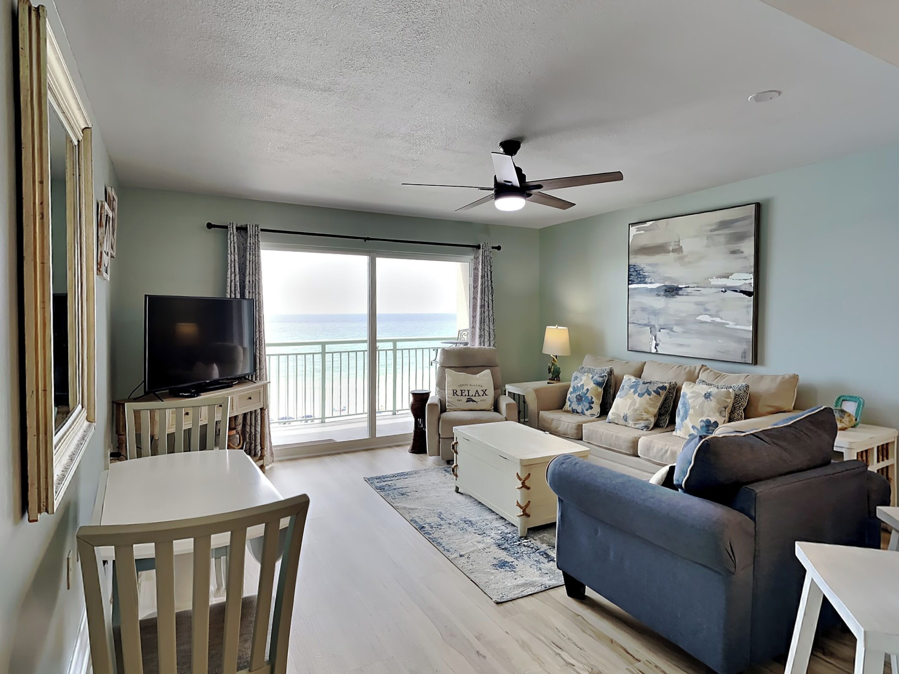 A living room with a TV, a ceiling fan, a sofa, a coffee table, and a view of the ocean through a sliding door.