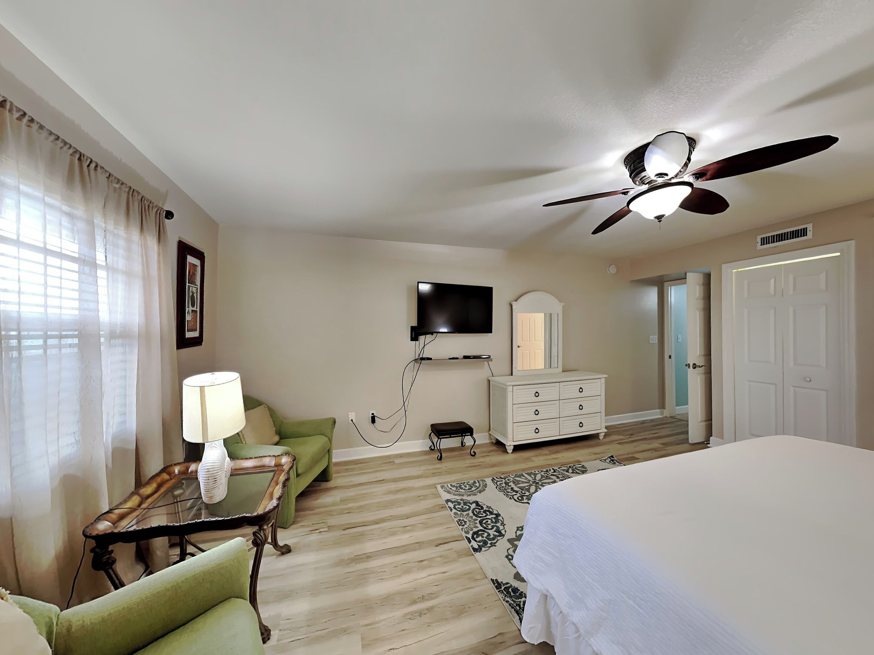 A hotel room with a bed, a desk with a lamp, a television mounted on the wall, a dresser, and a ceiling fan.