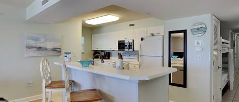 A modern kitchen with white cabinets, a central island with stools, a microwave, a refrigerator, and a clock on the wall.