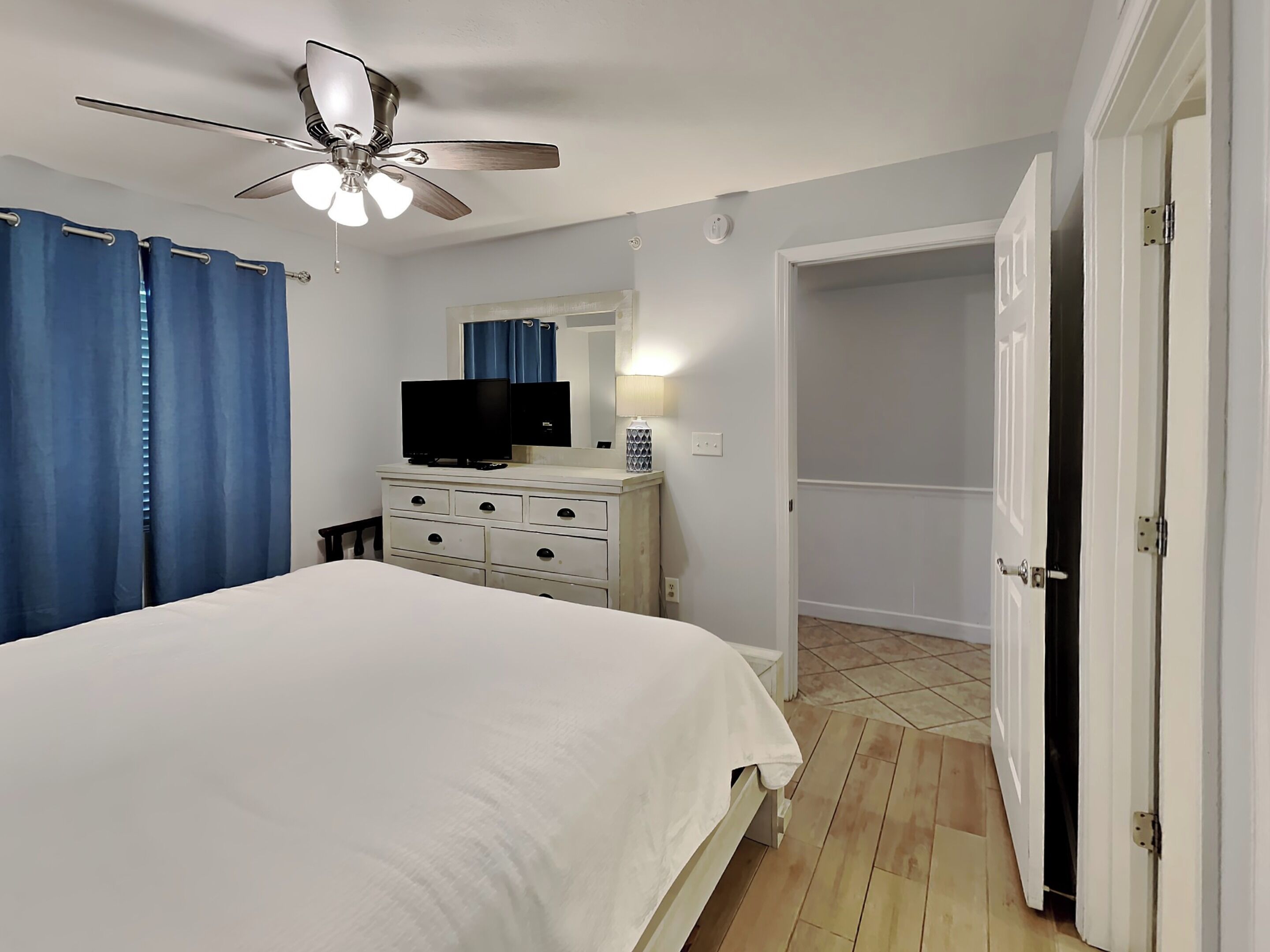 Un dormitorio con cama, cómoda, televisor, ventilador de techo y una puerta que da a otra habitación.