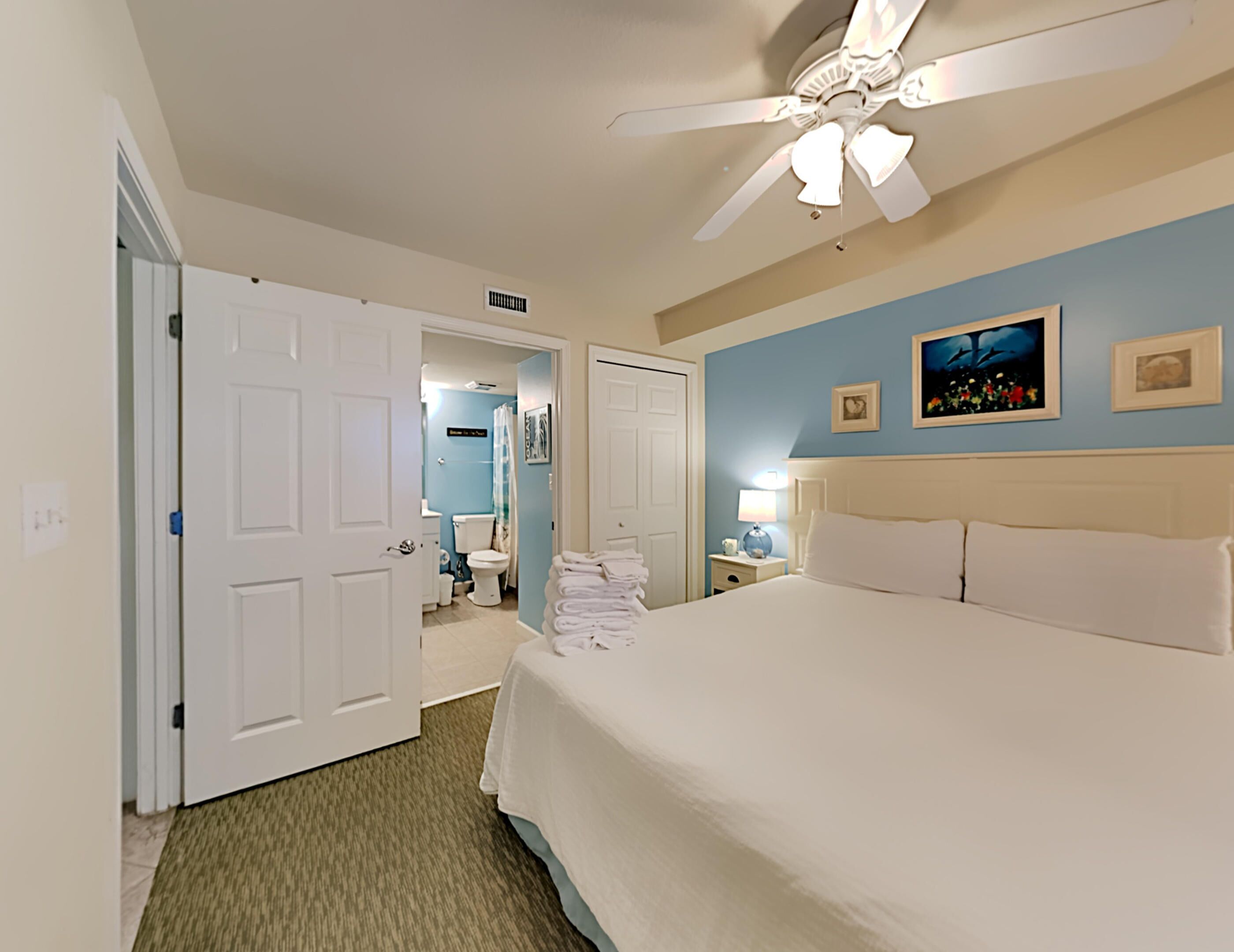 A hotel room with a large bed, a ceiling fan, a bathroom visible through an open door, and artwork on the walls.