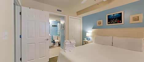 A hotel room with a large bed, a ceiling fan, a bathroom visible through an open door, and artwork on the walls.