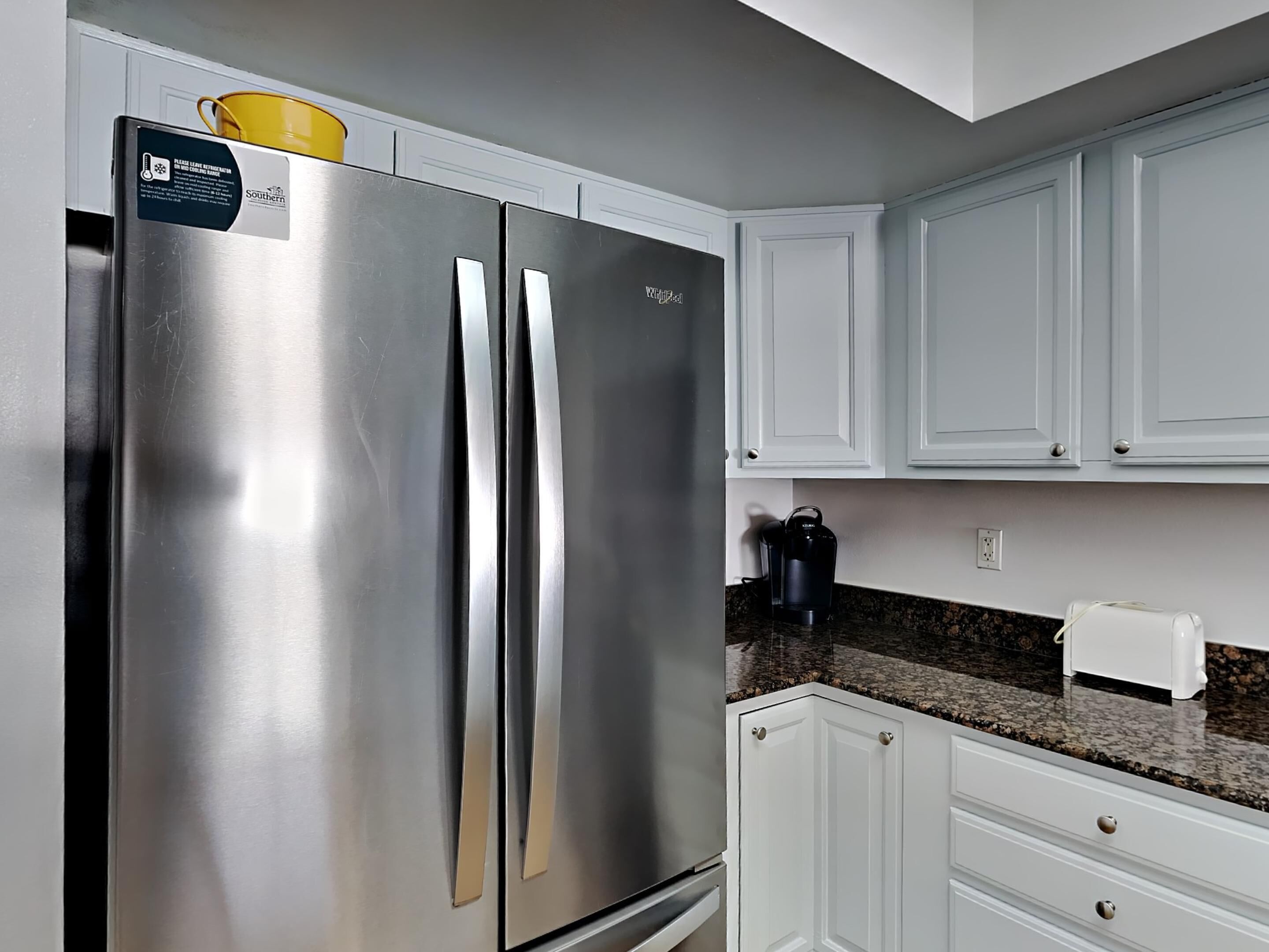 A stainless steel refrigerator with a yellow pot on top, white cabinets, and a coffee maker on the countertop.