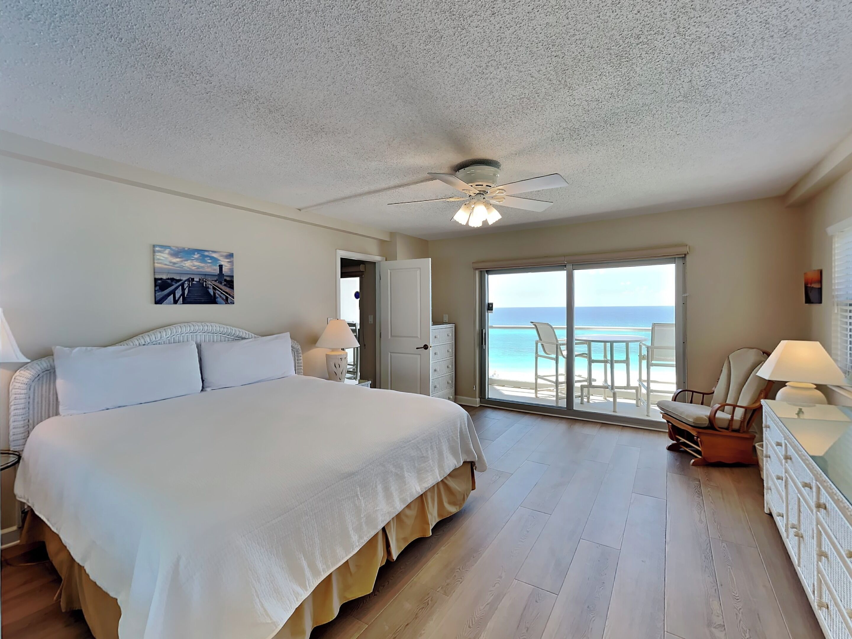 A spacious bedroom with a large bed, a ceiling fan, and a balcony with a view of the ocean.
