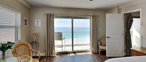 A bedroom with a view of the beach, a ceiling fan, a bed, and a dining area with wicker chairs.